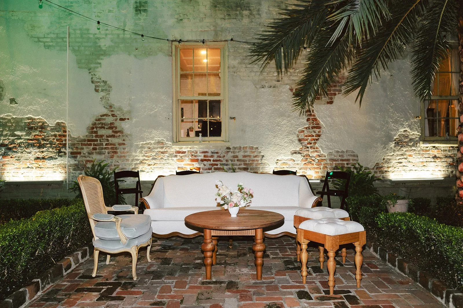 An outdoor patio with vintage-style furniture including a white sofa, a round wooden table with a floral arrangement, a light blue armchair, and small wooden stools. There are three chairs against a weathered brick wall with two windows, some potted 