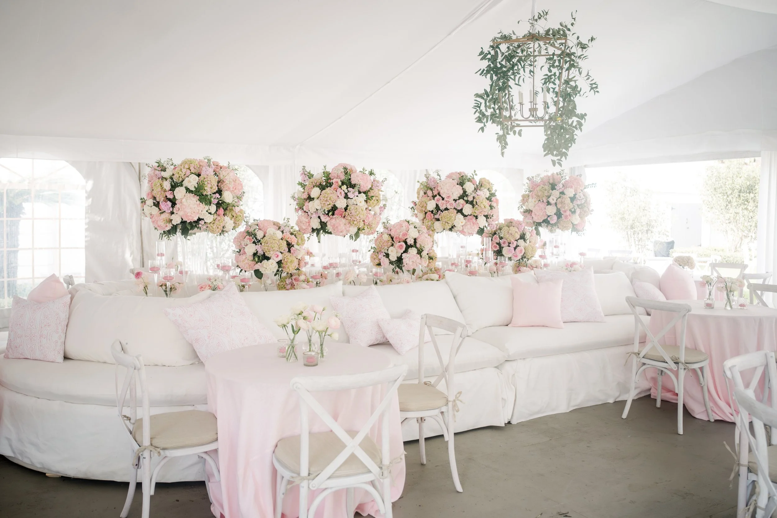 Elegant event setup inside a white marquee tent with pink and white floral arrangements on tables, white chairs with pink sashes, and a decorative hanging chandelier with greenery.