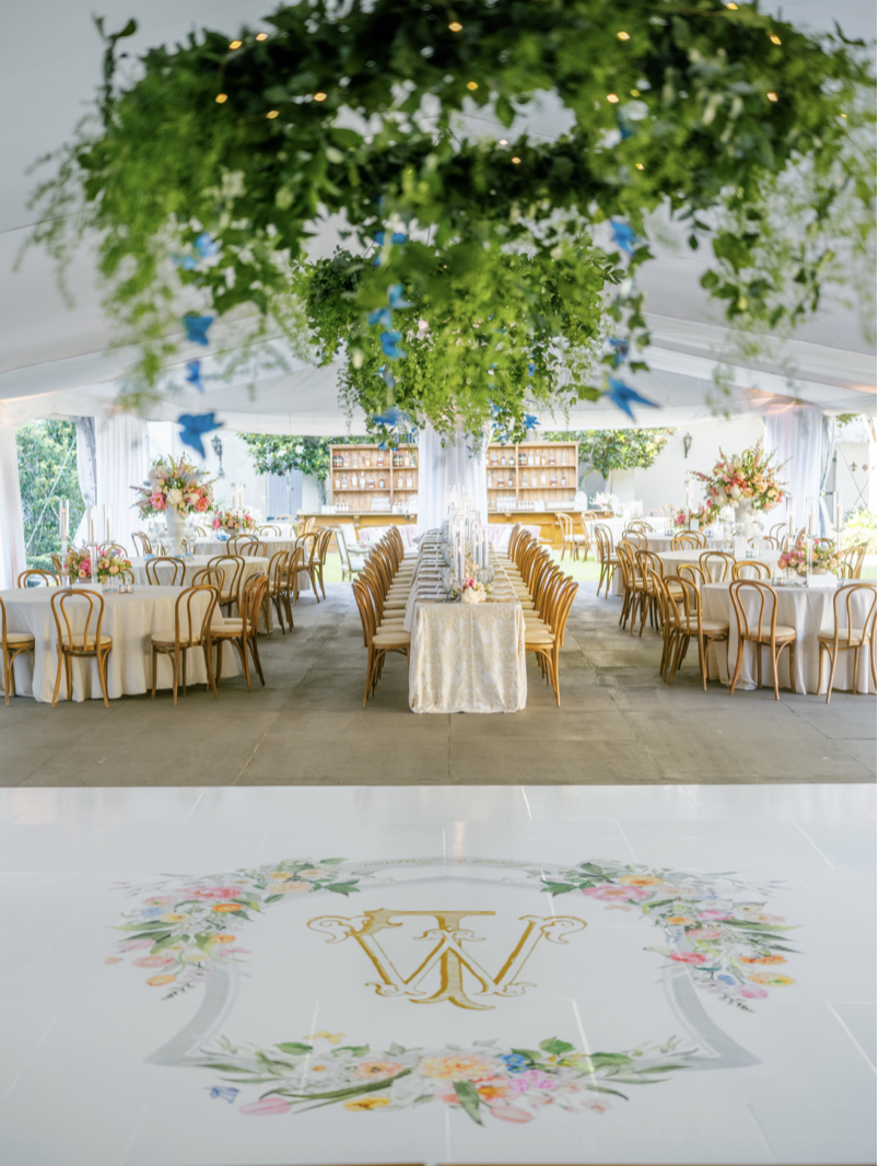 Elegant wedding reception tent with round tables, floral centerpieces, and hanging greenery with blue accents. A monogrammed initial 'W' is on the decorated floor at the entrance.