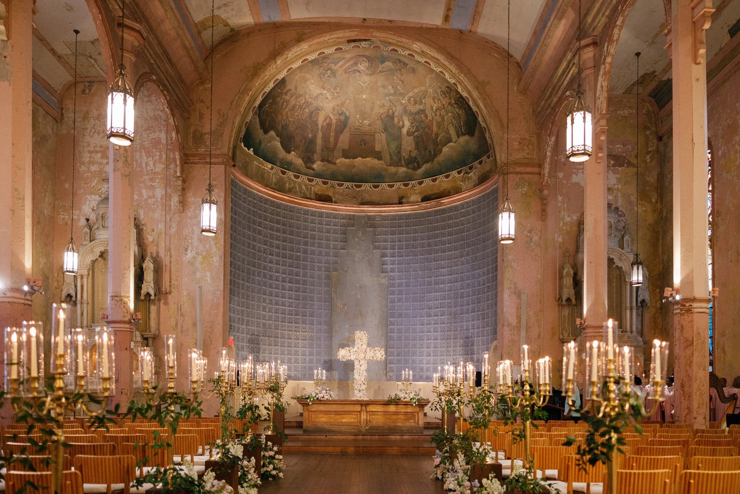 The interior of a church decorated for a wedding, with a large painted semicircular ceiling and rows of lit candles on either side of the aisle, leading up to an altar adorned with flowers.