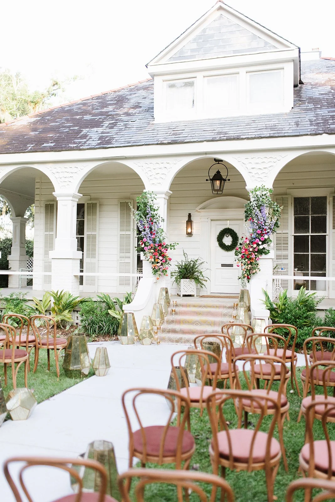 Front porch of a white house decorated for an outdoor event with pink and purple flowers, chairs, and lanterns.