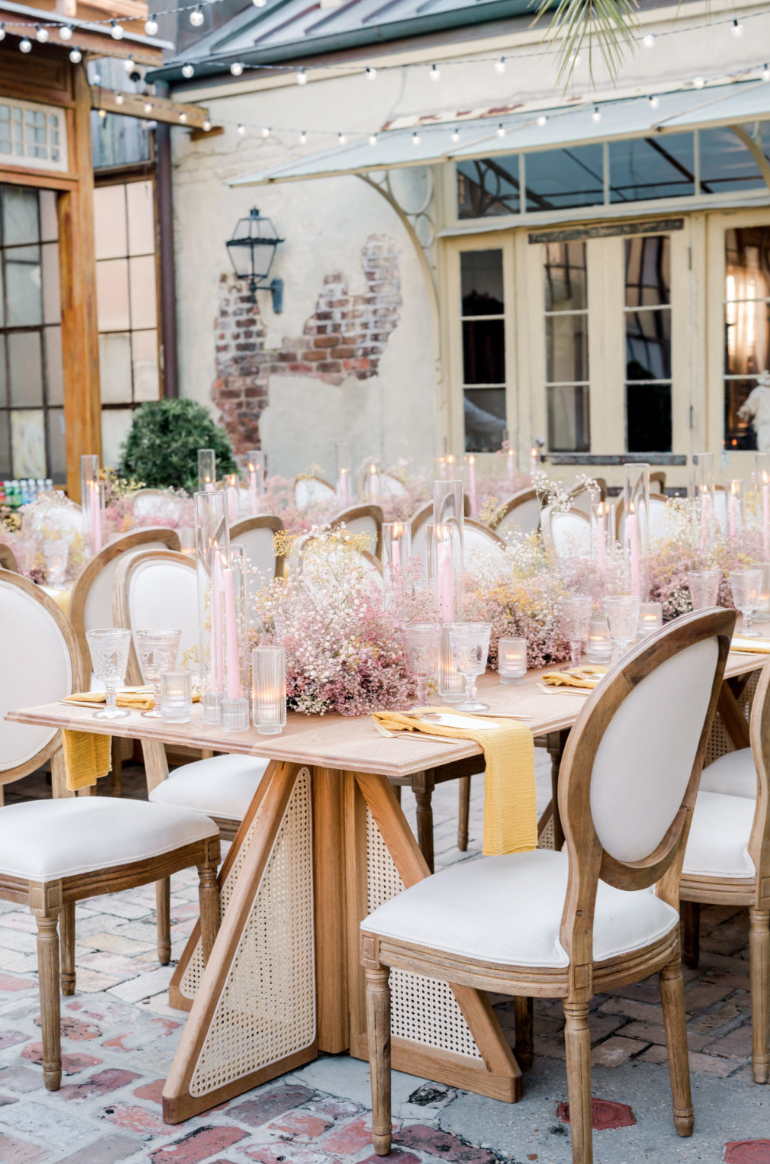 A decorated outdoor dining area with a long wooden table set for a celebration, featuring pink and white floral centerpieces, tall pink candles, glassware, and gold-accented plates, surrounded by white upholstered chairs with wooden frames, under str