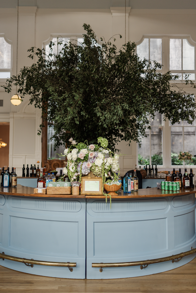 A bar with a large leafy plant behind it, decorated with a floral arrangement of white and pastel roses. Bottles of alcohol and cans of Sprite are on the bar, with windows and white walls in the background.