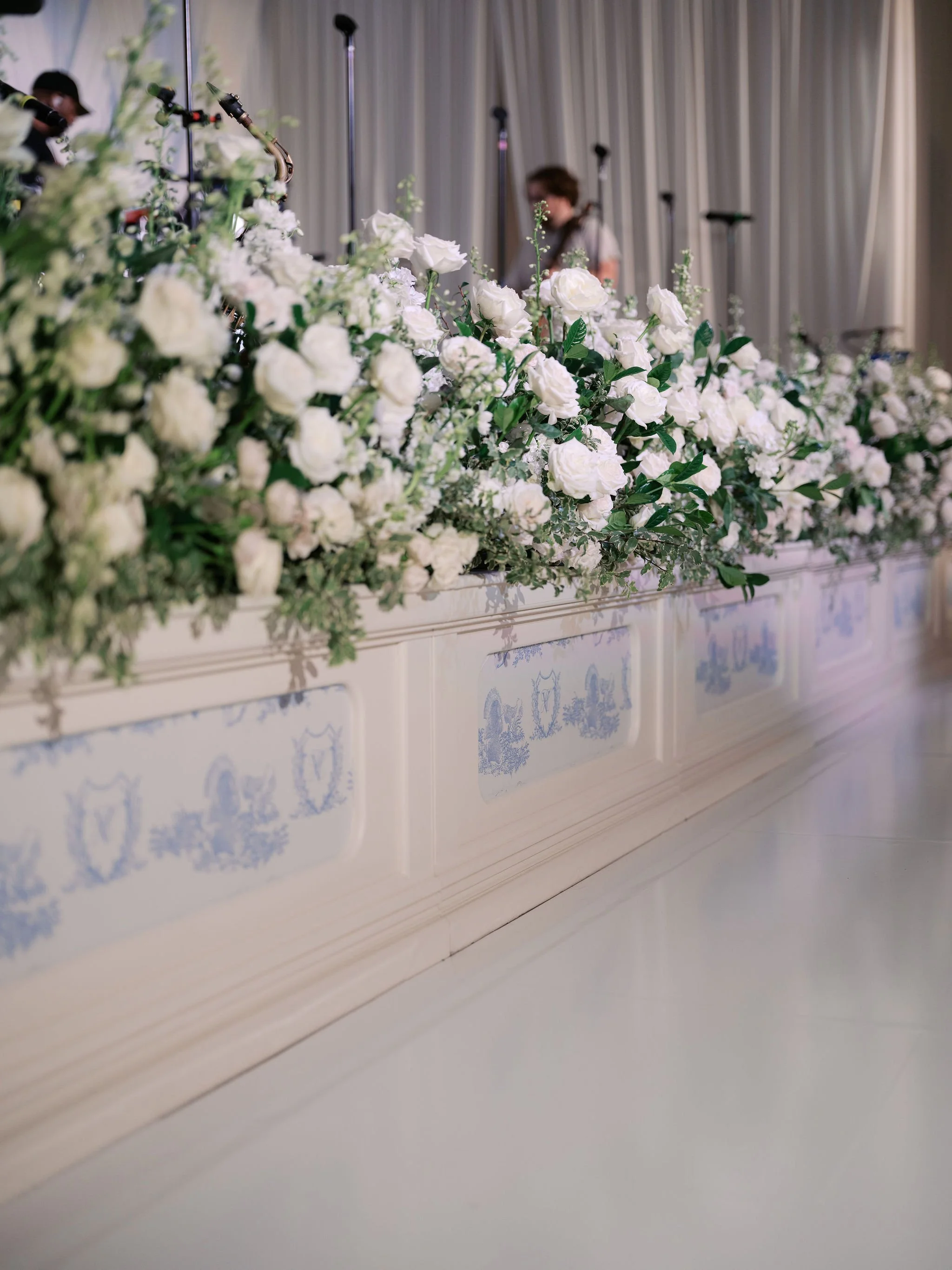 A row of white roses and greenery as a floral arrangement in front of a white decorated wall with blue pattern designs.