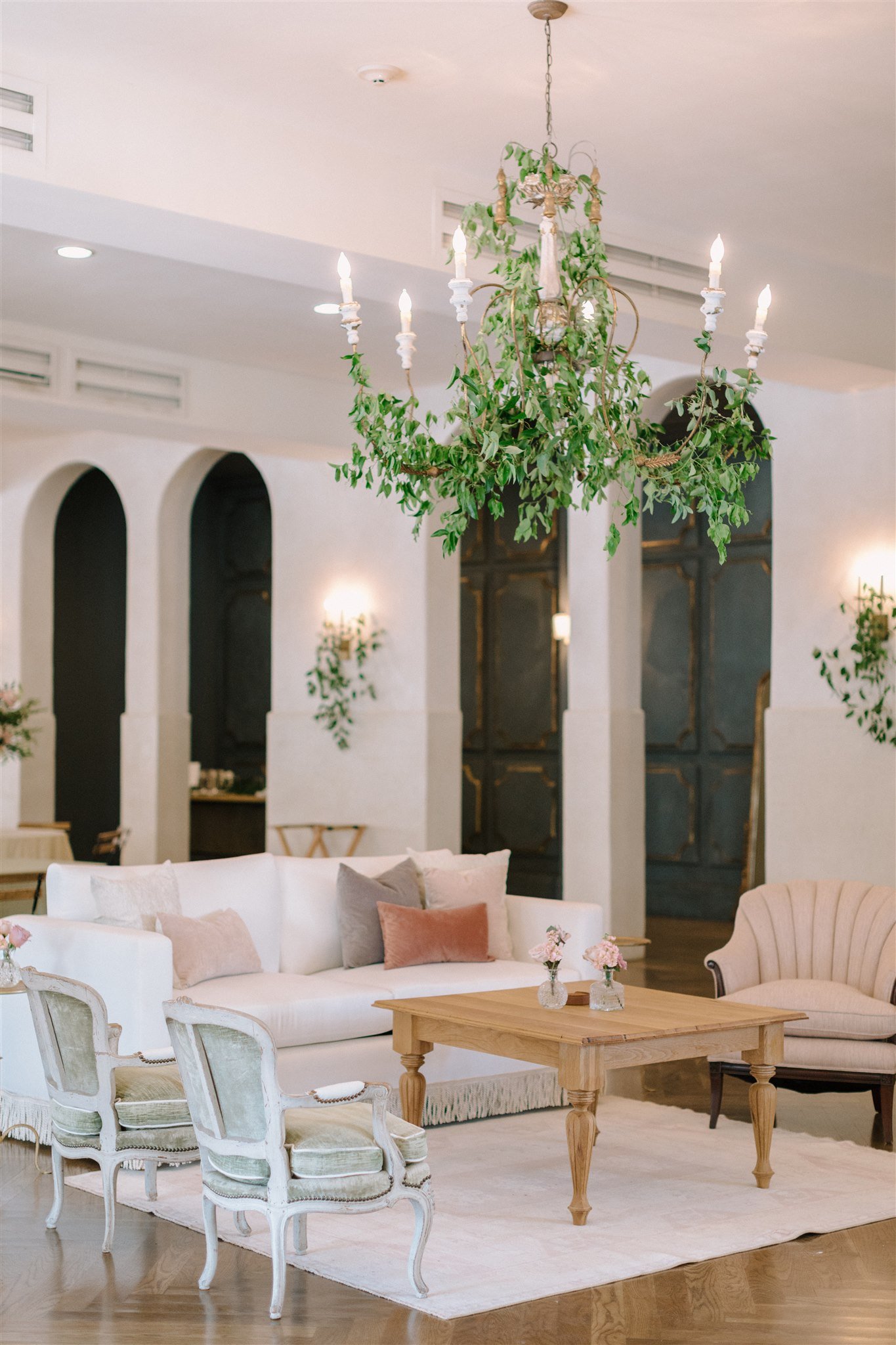 A living room with a chandelier decorated with green leaves. The room has white sofas, armchairs, a wooden coffee table with vases of pink flowers, and a light-colored rug. The background features arched wall niches and a dark door.
