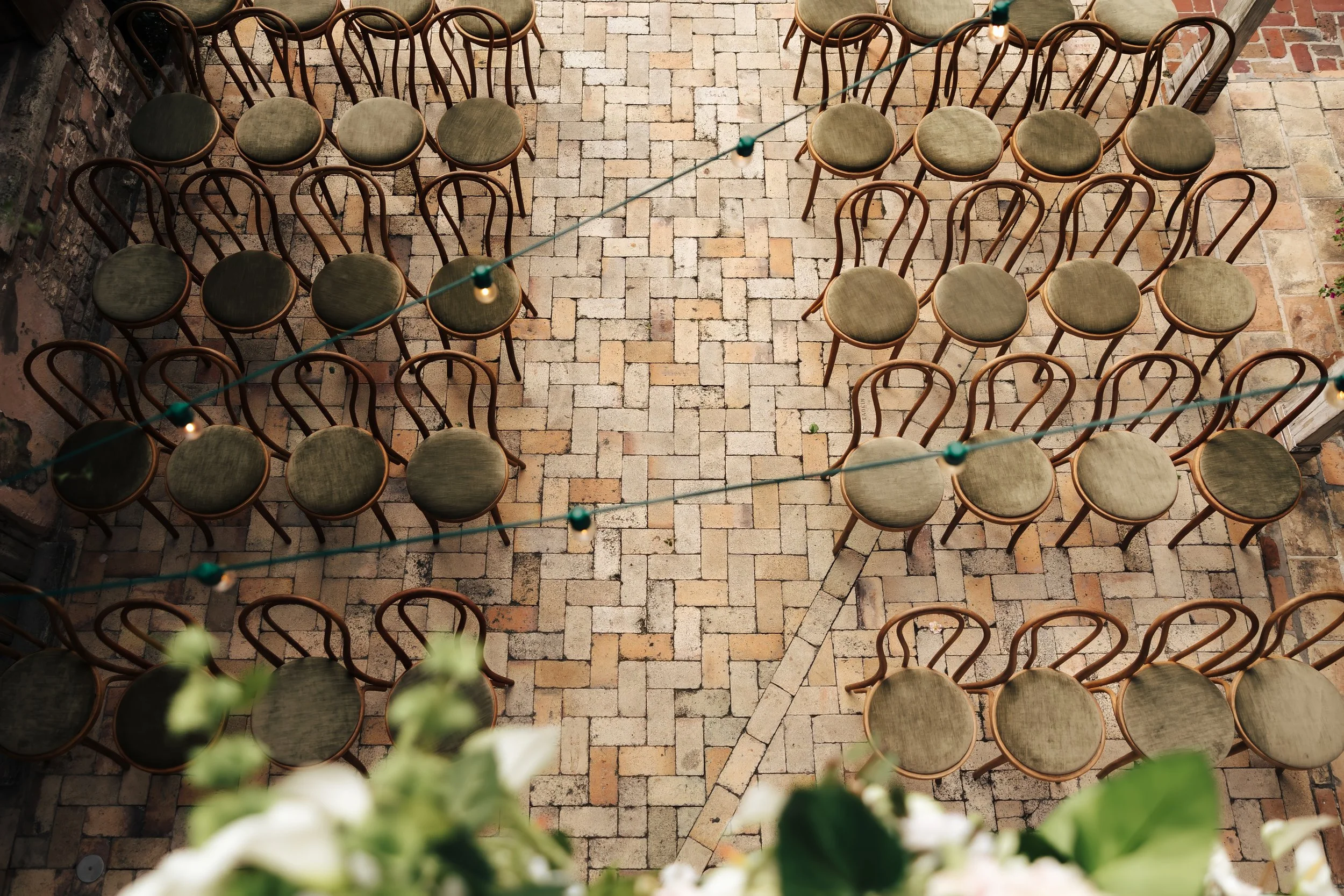 Empty green-upholstered chairs arranged in rows on a brick patio, with string lights overhead.