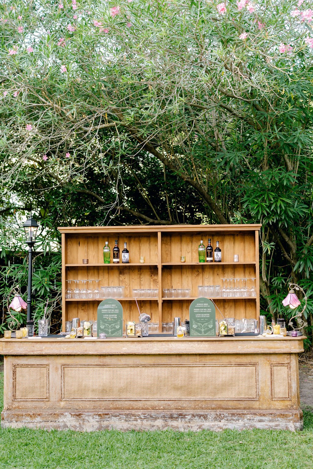 A rustic outdoor bar setup with bottles of liquor, glasses, and drink accessories on the wooden counter, set against lush green trees and pink flowering plants.