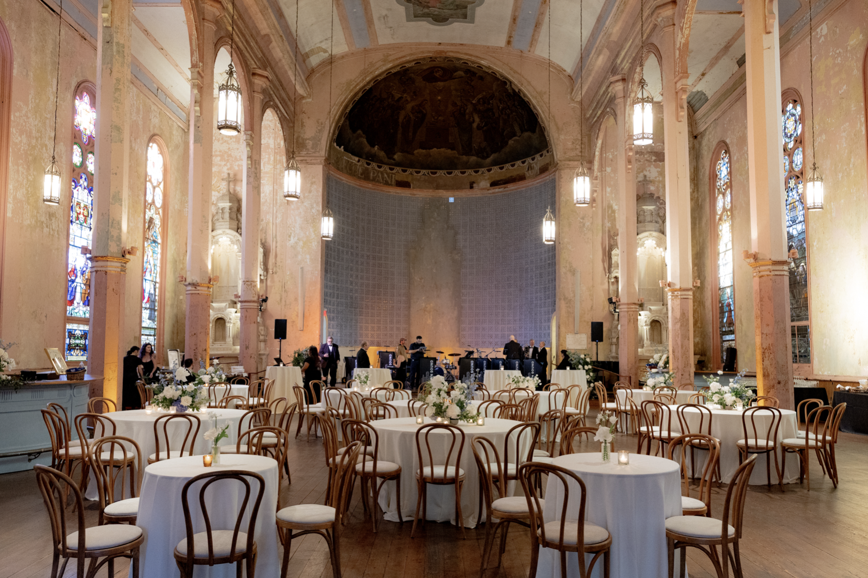 Elegant event space inside a historic church or chapel with high, arched ceilings, stained glass windows, and a stage where a band is setting up. Round tables with white tablecloths and floral centerpieces are arranged for guests, and small candles a