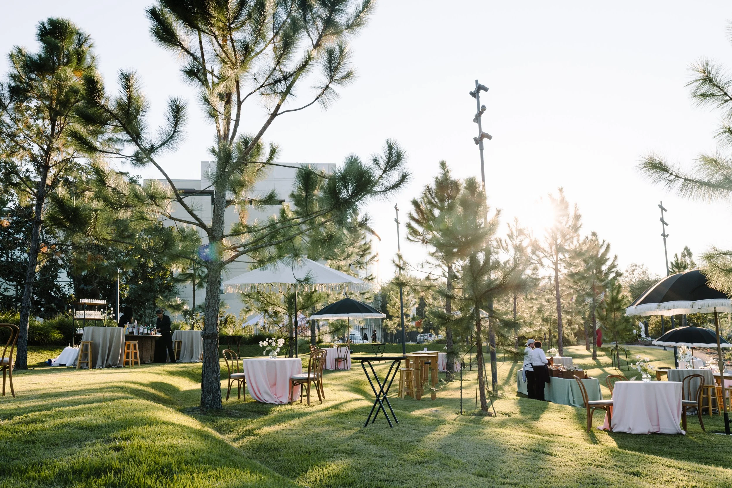 Outdoor event with round tables covered in pink and white tablecloths, surrounded by chairs, set on a grassy area with tall pine trees. The sun is shining brightly, and there are umbrella-covered seating areas and a bar with drinks being served.
