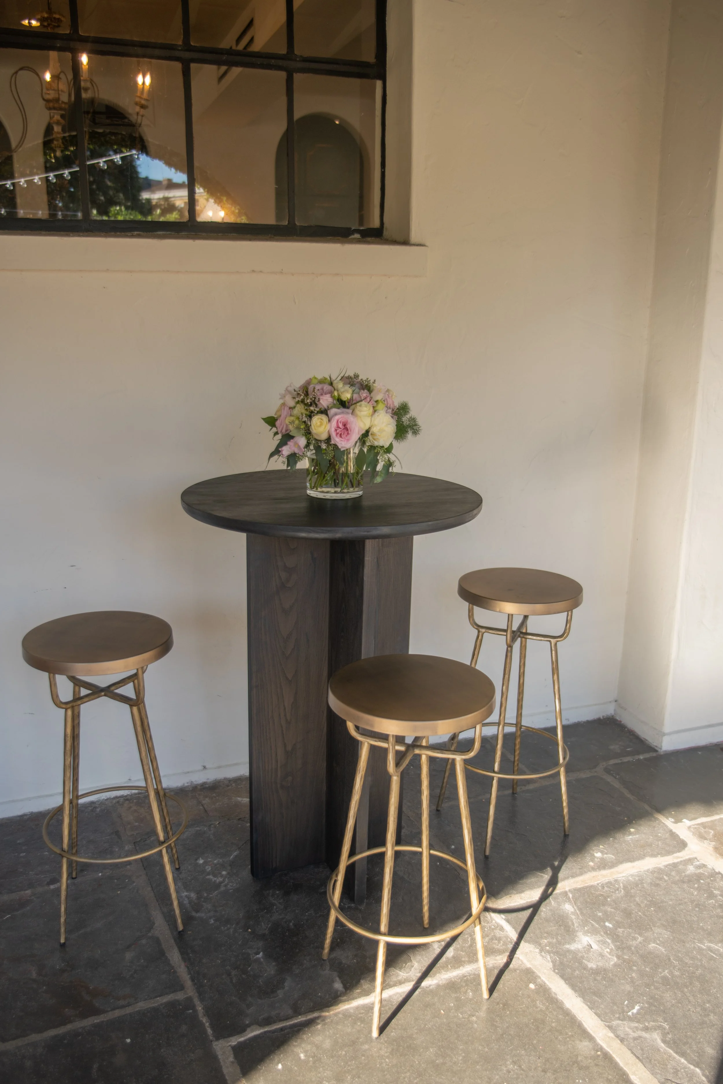 A round, dark wooden table with a glass vase of white, pink, and purple flowers. Four gold-colored metal bar stools with round wooden seats are placed around the table. The setting appears to be in a cozy, indoor-outdoor space with natural light, off