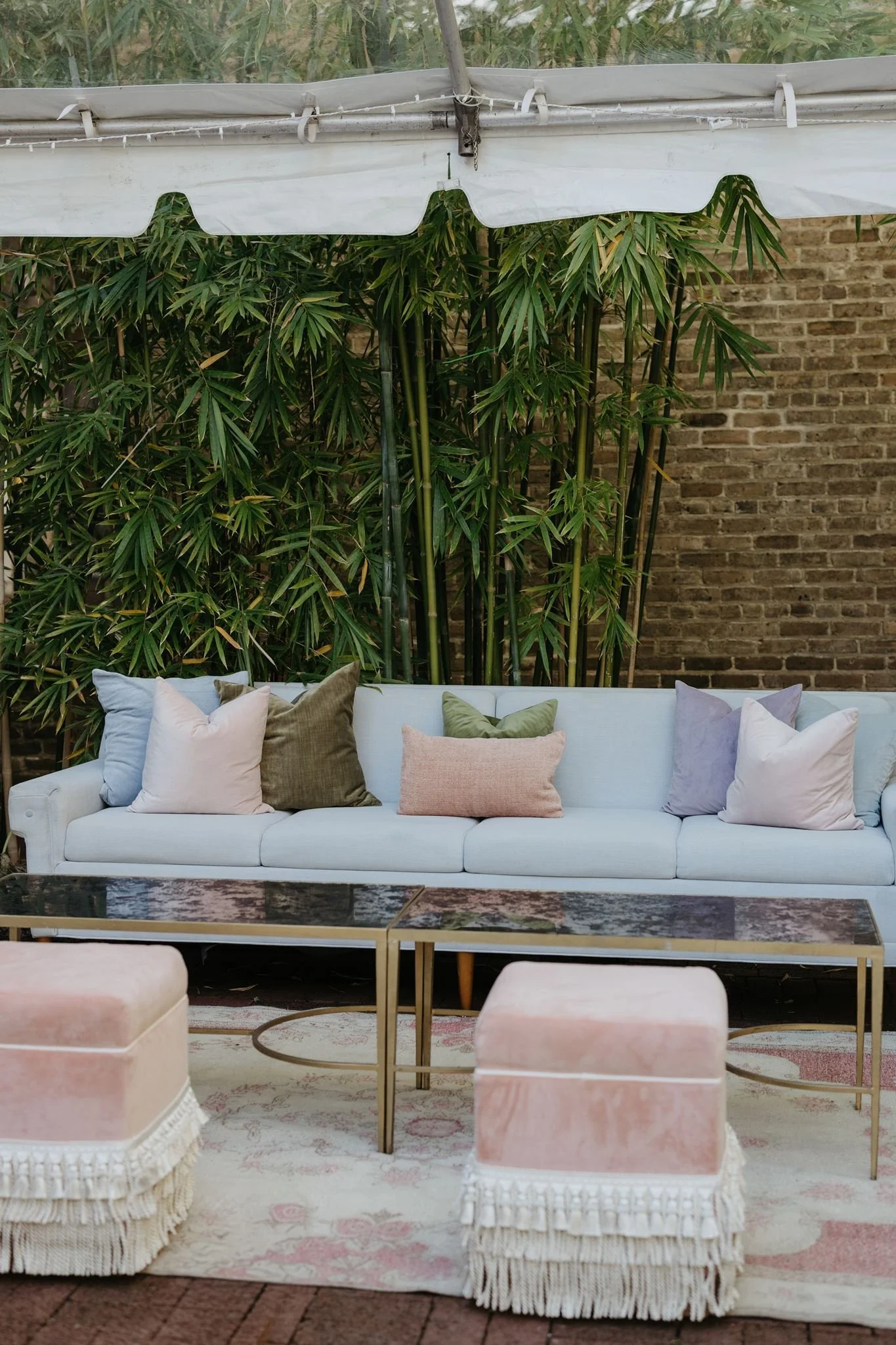 A white sofa with multiple pastel-colored throw pillows on a patterned rug. In front of the sofa, there are two pink ottomans with fringed skirts, and a glass-top coffee table with a gold frame. Behind the sofa, there is a brick wall and tall green p