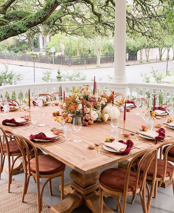 A tablescape set on a wooden table with a floral centerpiece, surrounded by chairs, on a porch with a view of a street and trees.