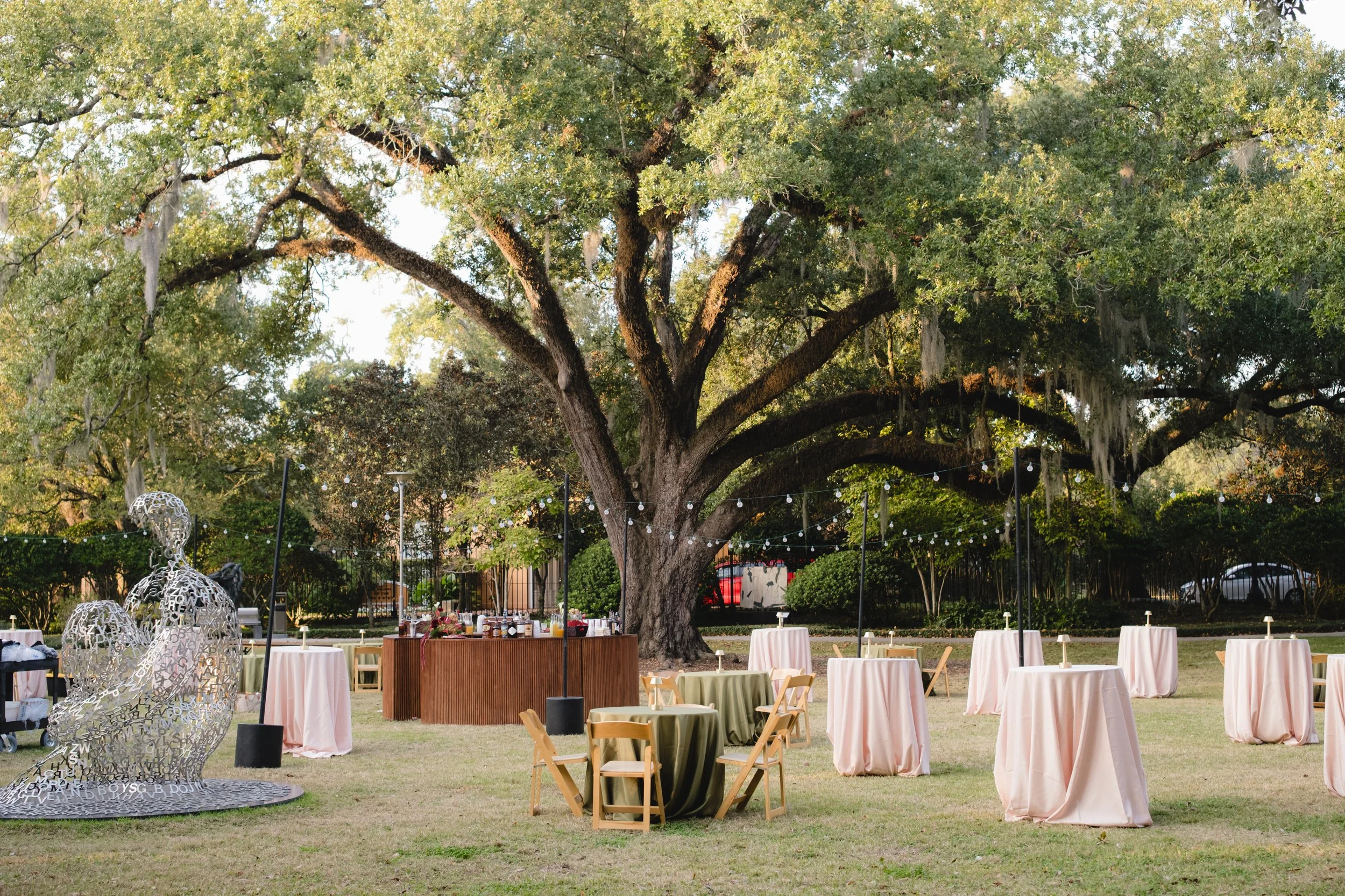 Outdoor event setup with round tables covered in pink and green tablecloths, surrounded by chairs, under large leafy trees. Decorative hanging lights and abstract art sculptures are also visible.