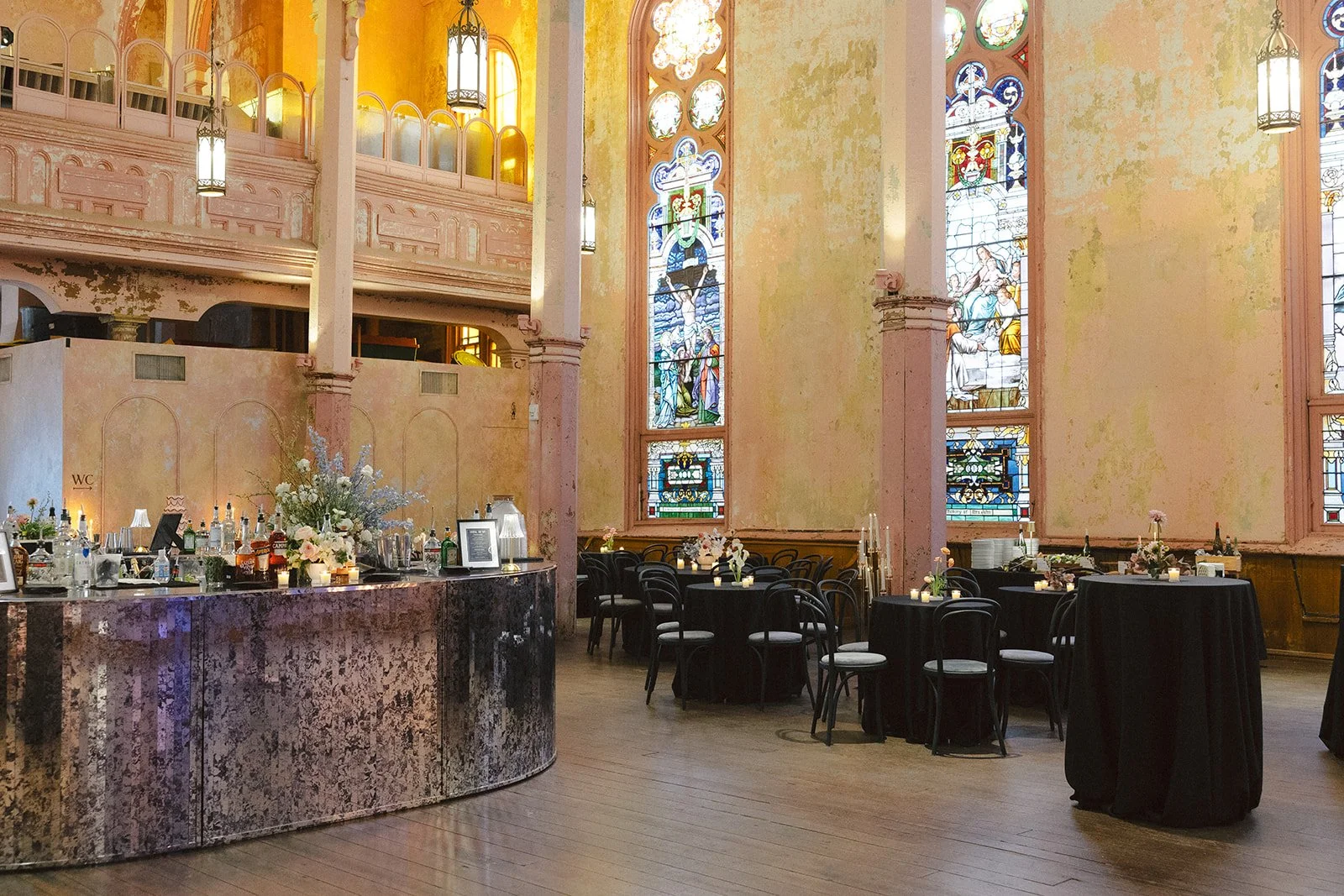 Interior of a decorated event space with stained glass windows, a bar area, and round tables with floral centerpieces and candles.