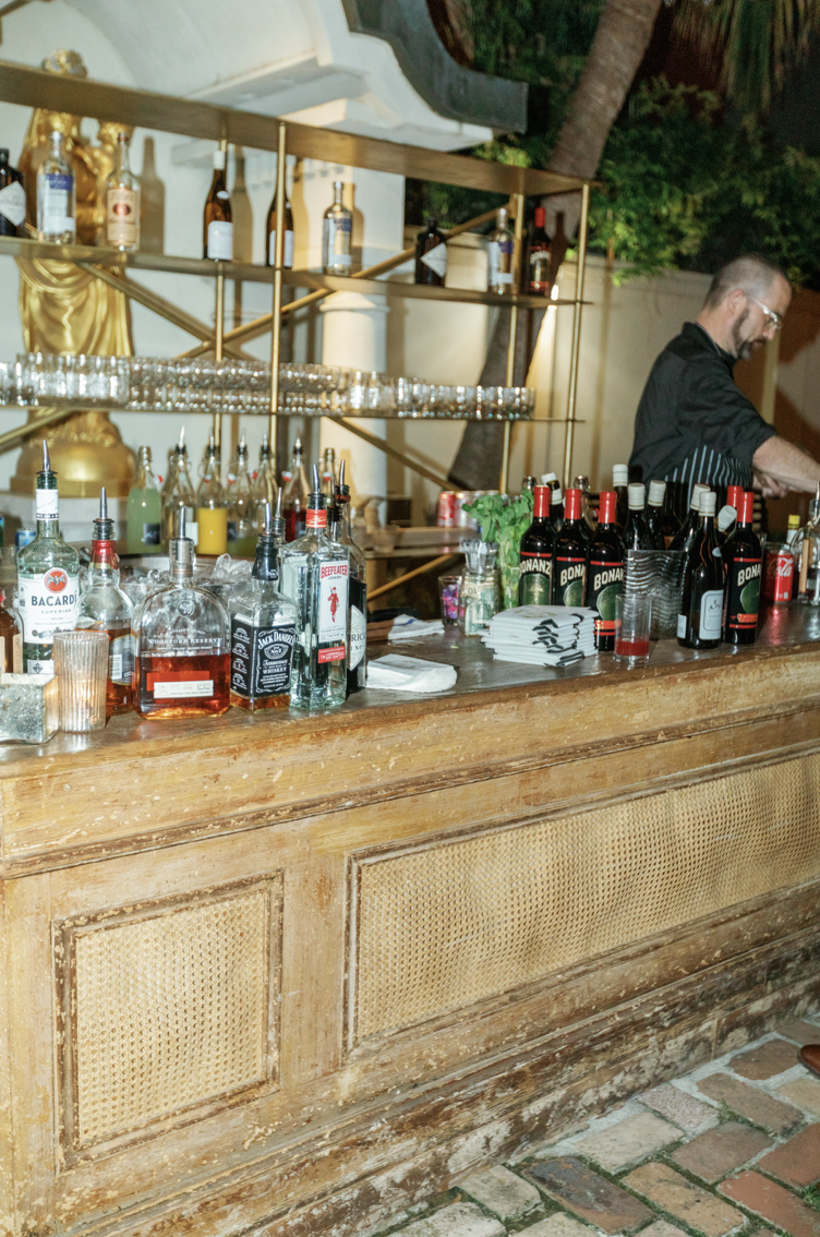 Bar with various liquor bottles, shot glasses, and a bartender preparing drinks at an outdoor venue at night.