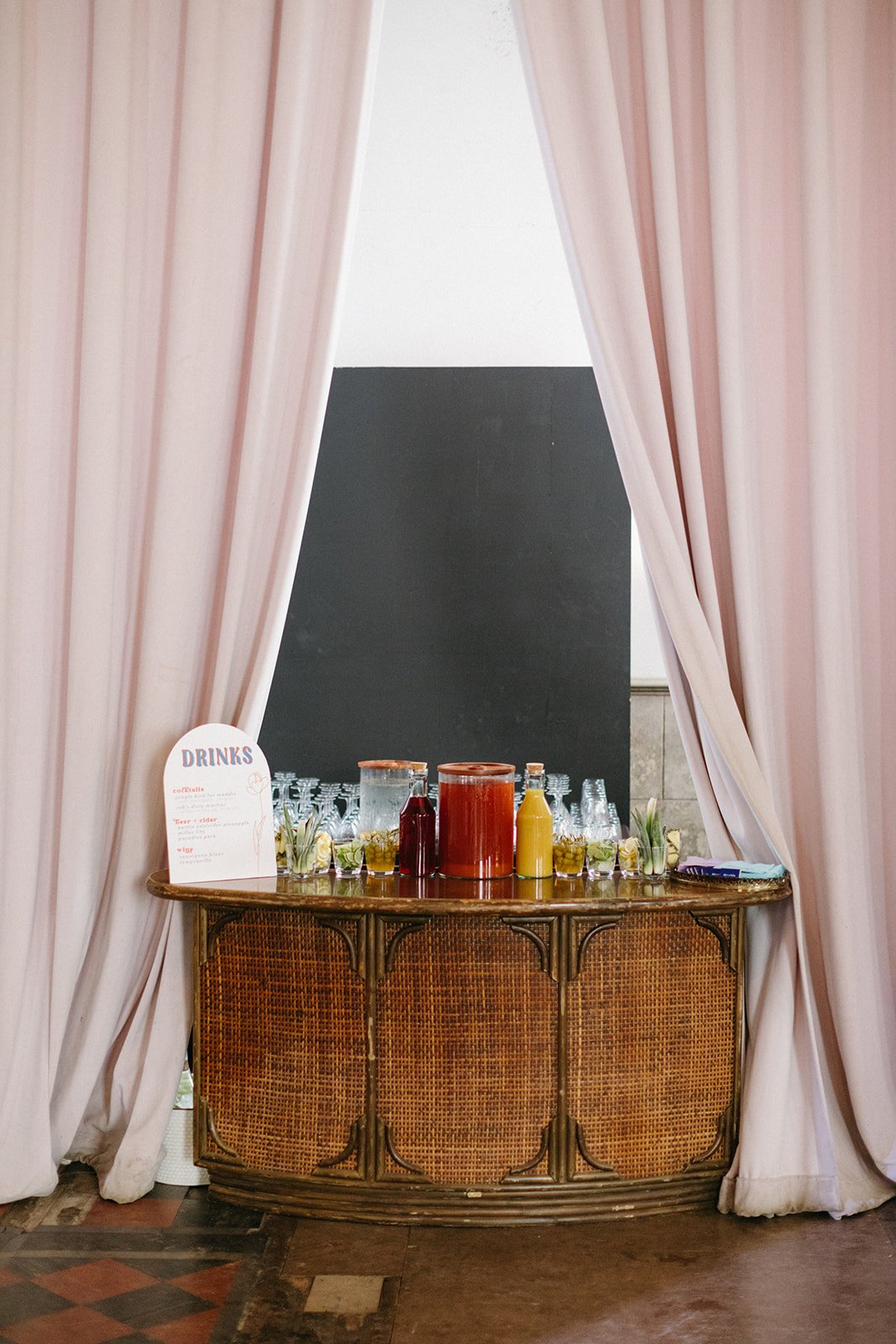 A vintage wicker bar cart behind pink curtains with a drink menu sign, glass pitchers, and condiment bottles.