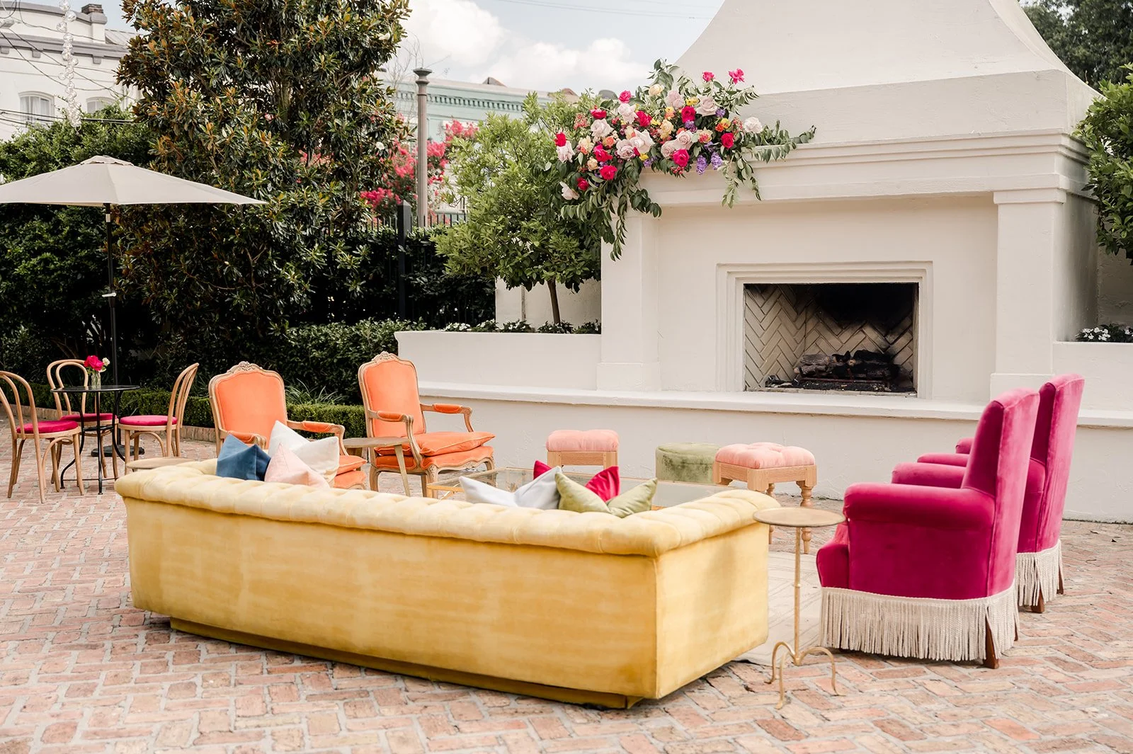 Colorful outdoor seating area with a yellow sofa, pink armchairs, and a small black table with pink chairs on a brick patio, adjacent to a white fireplace with a floral arrangement on top, surrounded by greenery.