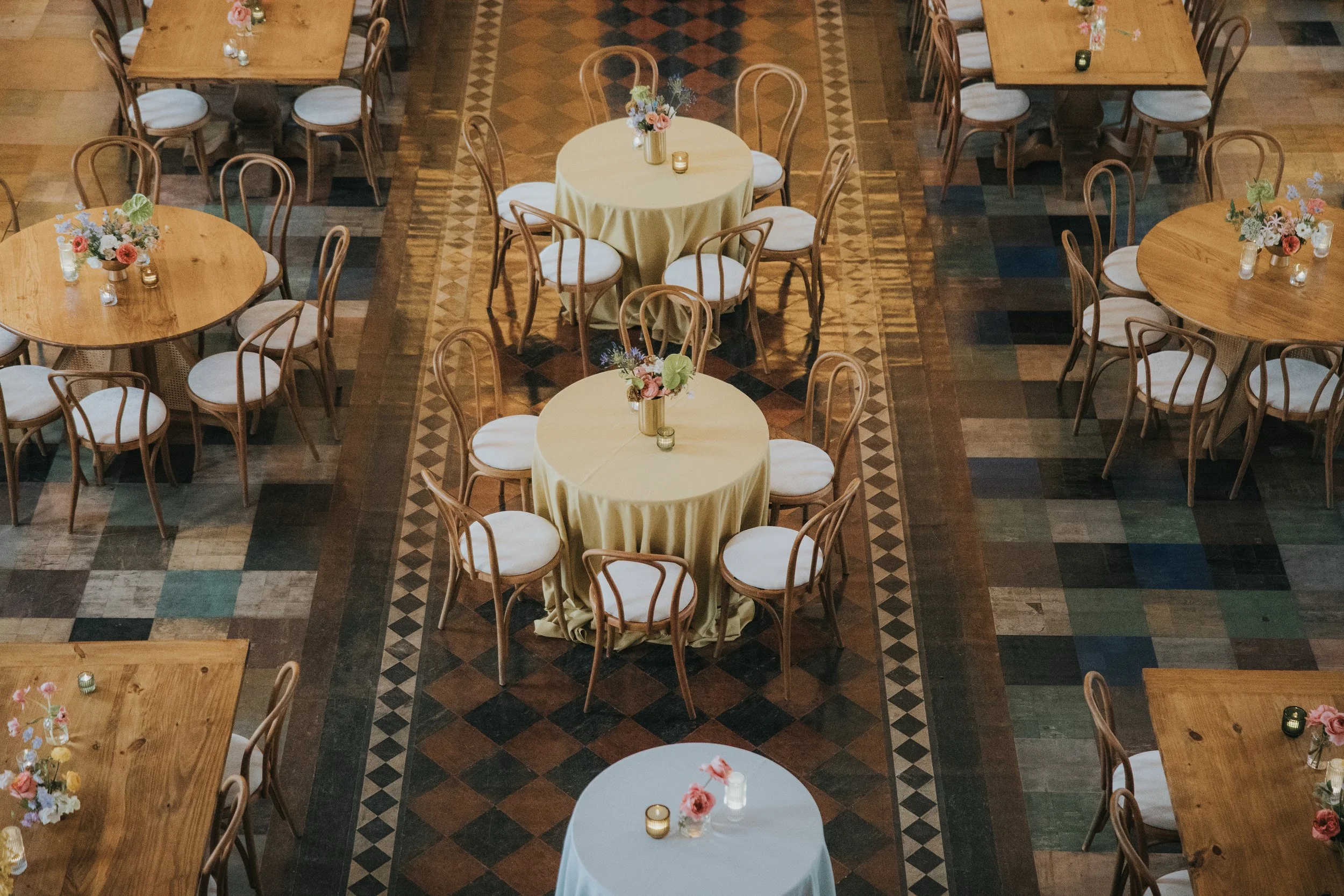 Empty event space with round tables decorated with floral centerpieces, candles, and white chairs, featuring patterned wooden flooring.