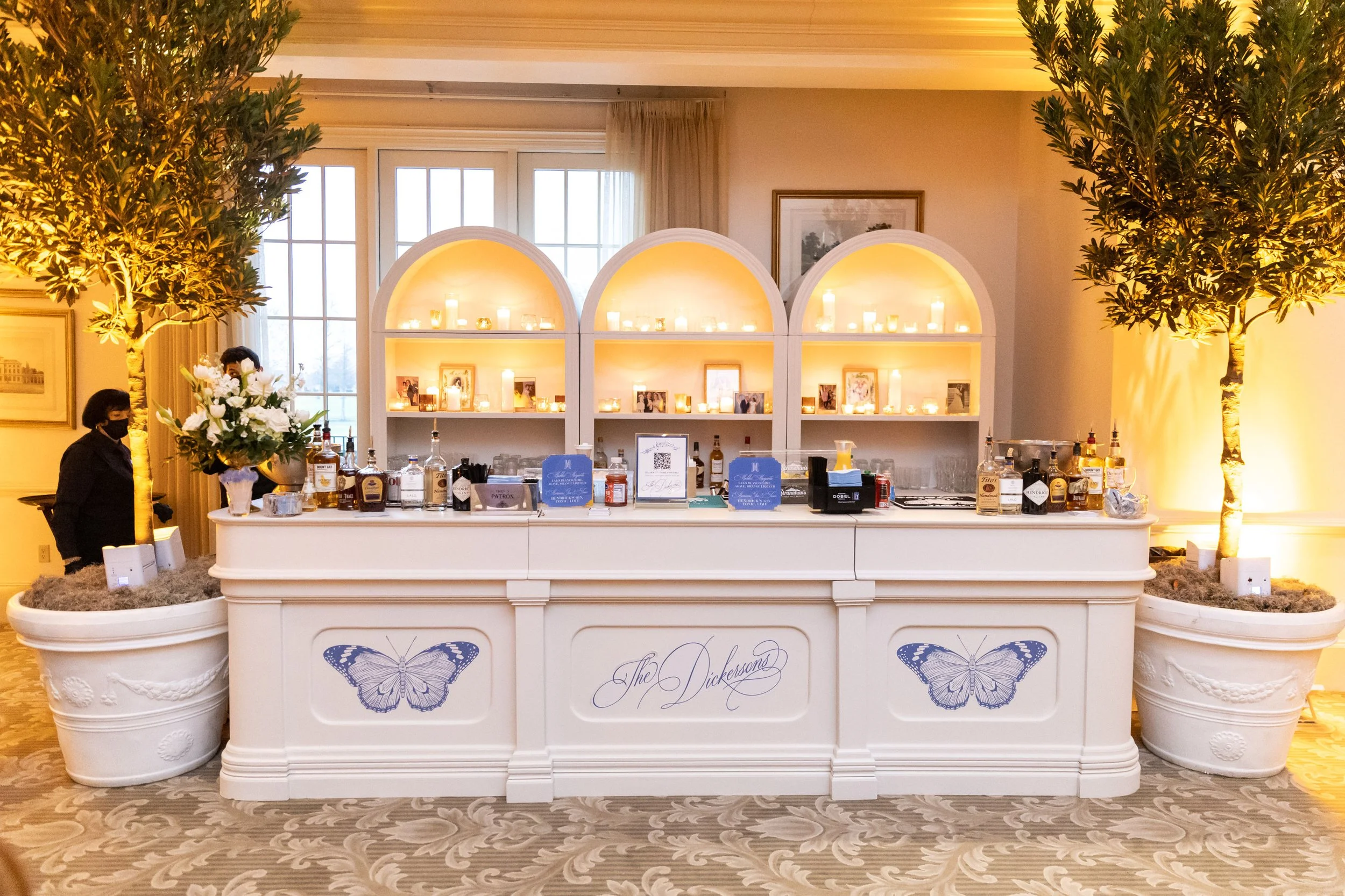 A white bar with two large potted plants on either side, decorated with butterfly and floral motifs. Behind the bar are three illuminated arches with candles, framed photos, and bottles of alcohol, set against a large window and framed art on the wal