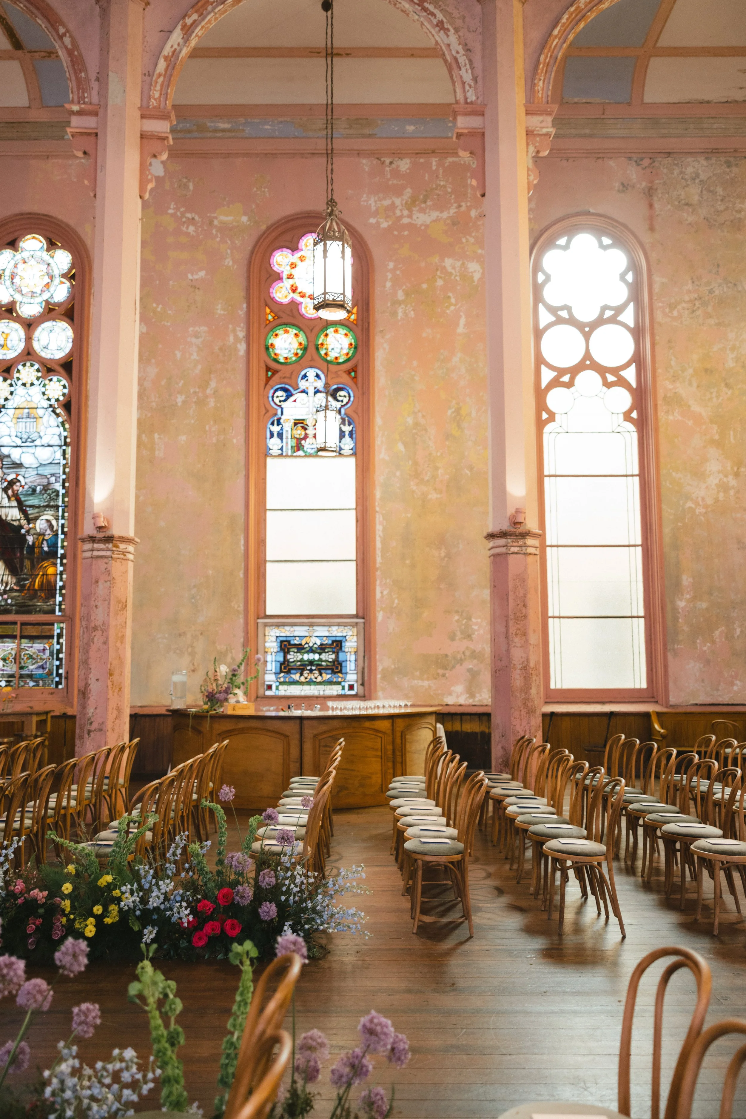 An interior view of an old church with worn pink walls, large stained glass windows, and wooden chairs arranged for a ceremony, with flower arrangements in the foreground.