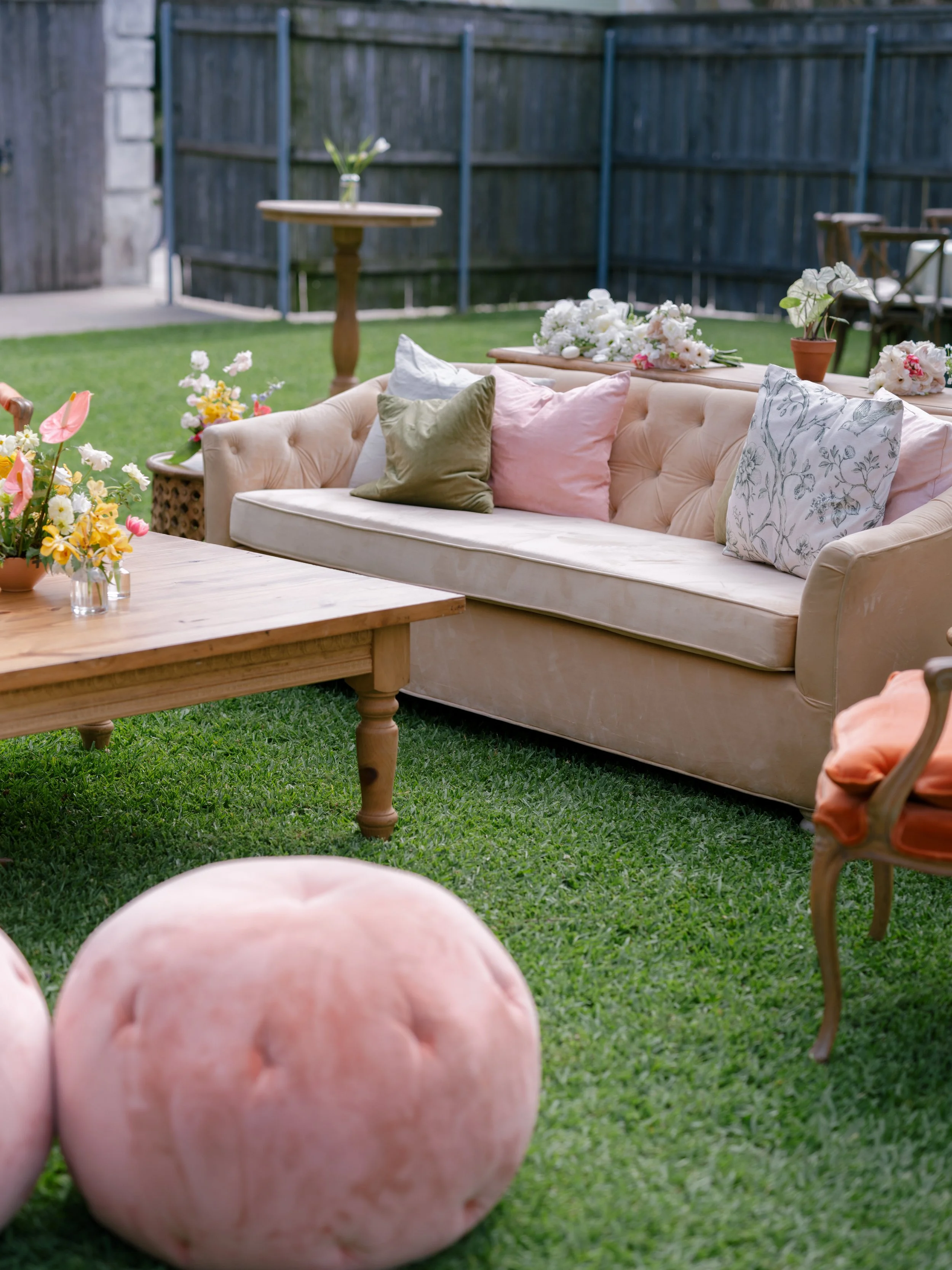 A backyard scene with a beige tufted sofa adorned with pastel-colored pillows. There are potted plants and flowers on and around the sofa. In the foreground, a large pinkish-orange ball is on green grass, with a wooden table and a wooden chair nearby