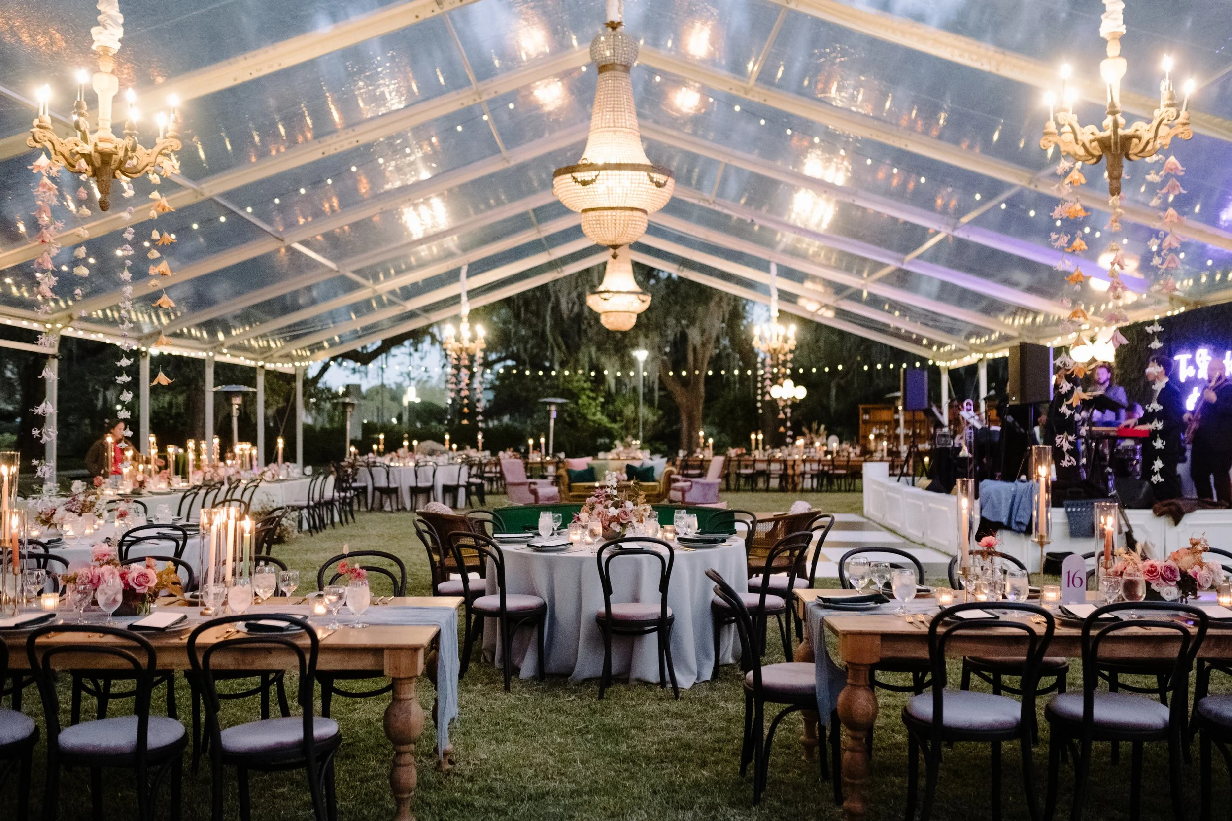 An elegant outdoor event space with round tables decorated with pink floral arrangements, candles, and glassware, under a transparent tent with chandeliers and string lights hanging from the ceiling, and a band performing on stage in the background.