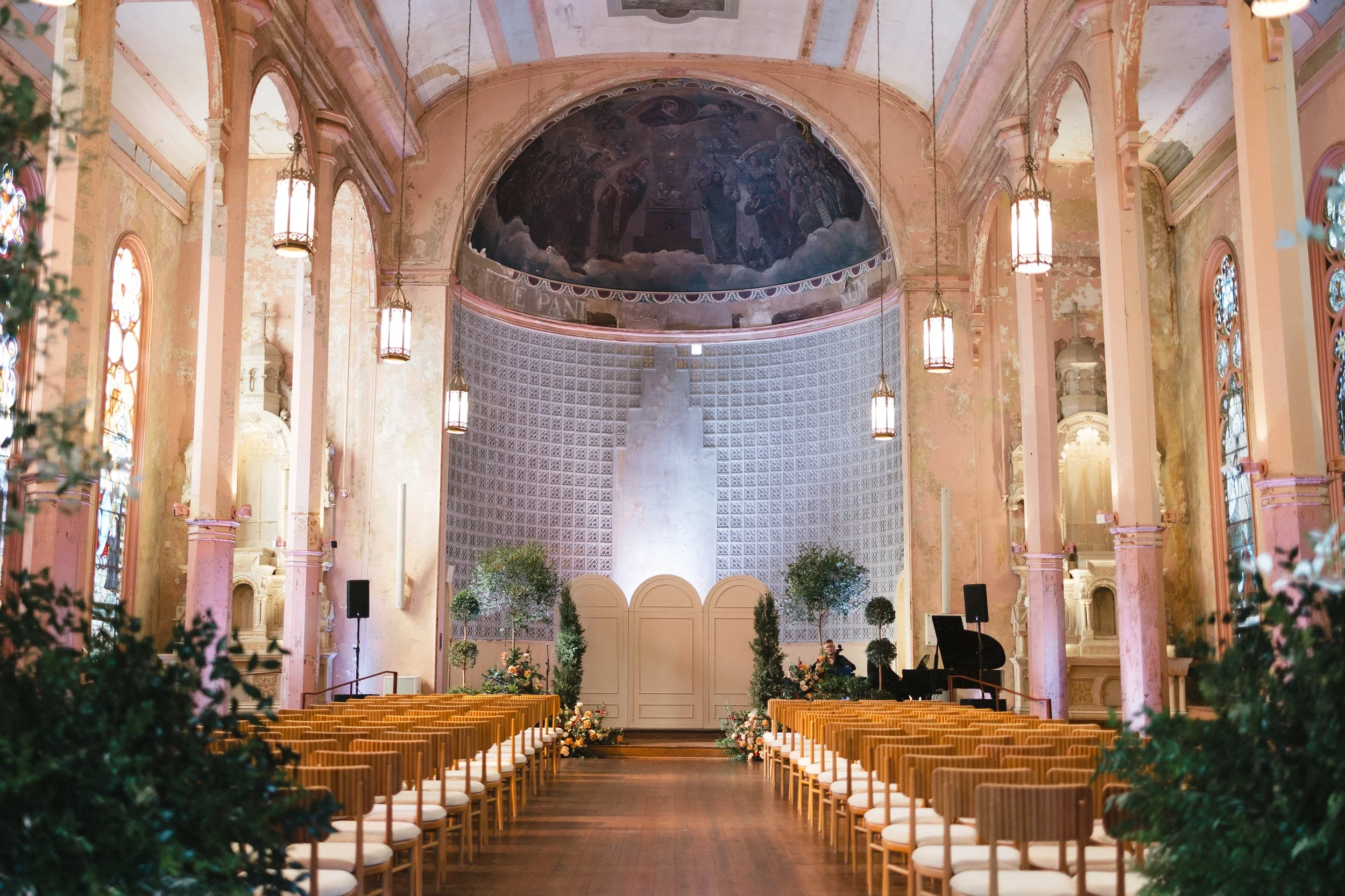 Interior of a church decorated for a wedding with rows of chairs, floral arrangements, potted trees, stained glass windows, hanging chandeliers, and a painted ceiling featuring religious imagery.
