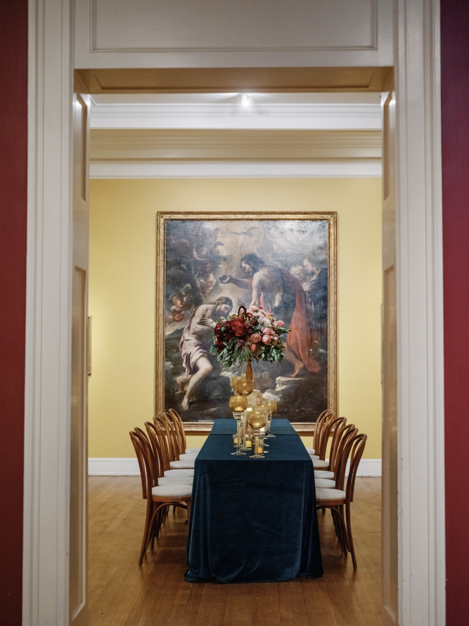 Elegant dining room with a long table covered with a dark blue tablecloth, surrounded by wooden chairs with white cushions. A tall floral centerpiece and gold-colored candlesticks decorate the table. The room features a large classical painting with 