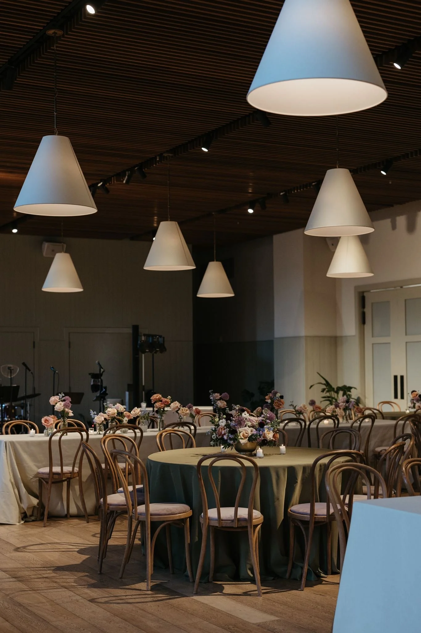 Interior of a event hall decorated for a celebration with long tables covered with white and dark green tablecloths, floral centerpieces, candles, and wooden chairs with padded seats. Ceiling has multiple white conical pendant lights and wooden panel