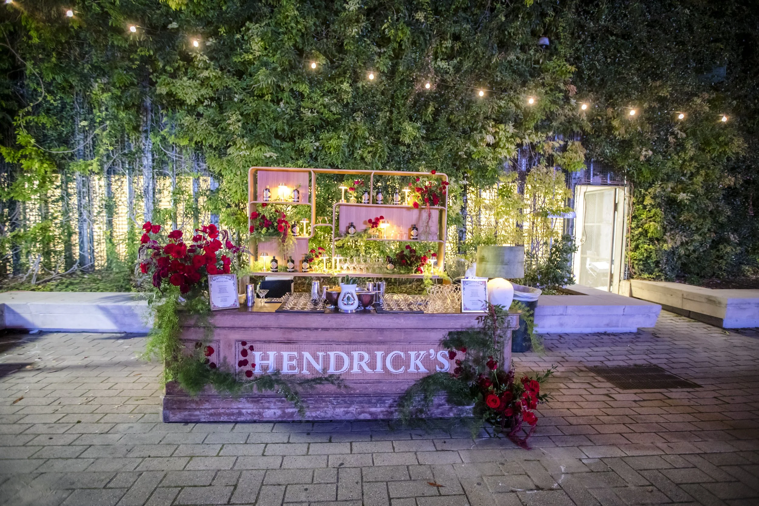 Outdoor event setup with a reception or bar area decorated with red flowers, greenery, and string lights hanging above, under a large tree, with a sign reading 'Hendrick's' on the front.