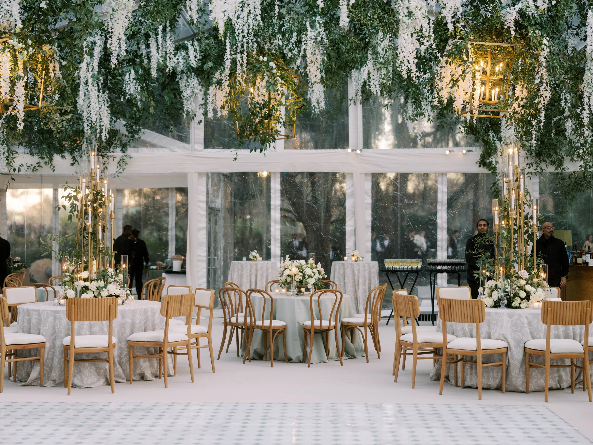 Elegant event space decorated with hanging white floral arrangements and gold lanterns, set with round tables covered in white tablecloths and surrounded by wooden chairs with white cushions."