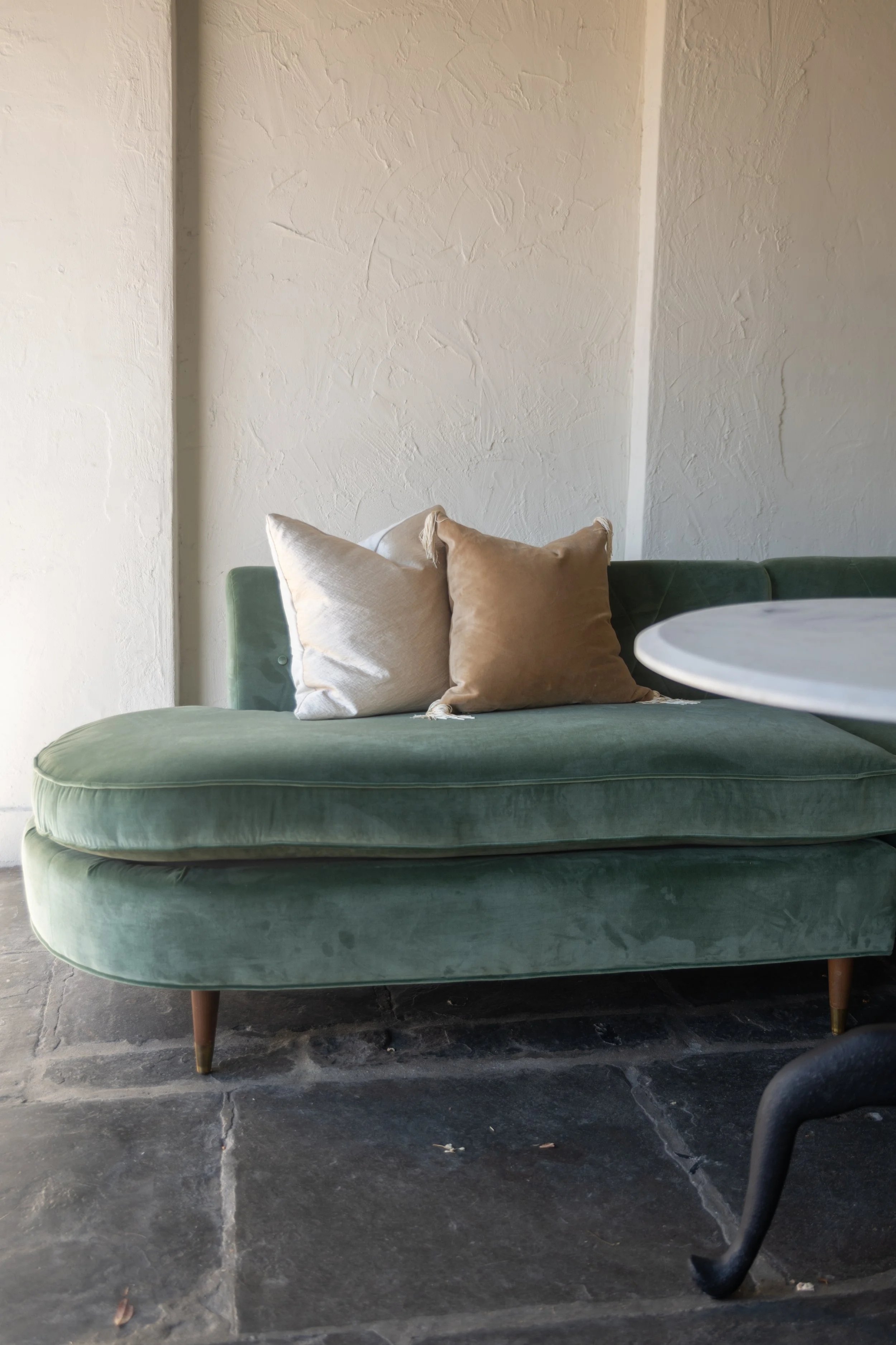 A green velvet mid-century modern sofa with wooden legs, topped with beige and light brown pillows, in front of a textured beige wall with a white round table partially visible in the foreground, on a dark stone floor.
