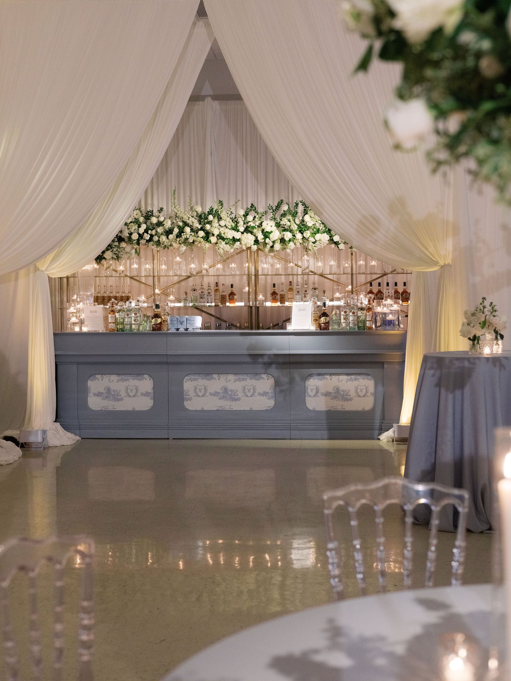 A bar area decorated with white flowers and candles, framed by cream-colored draped curtains, with a table and chairs in the foreground.