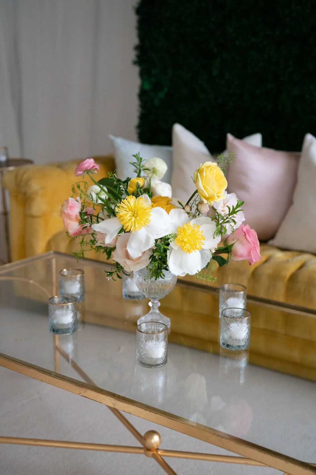 A floral centerpiece with yellow, white, and pink flowers on a glass coffee table, surrounded by small glass candleholders.