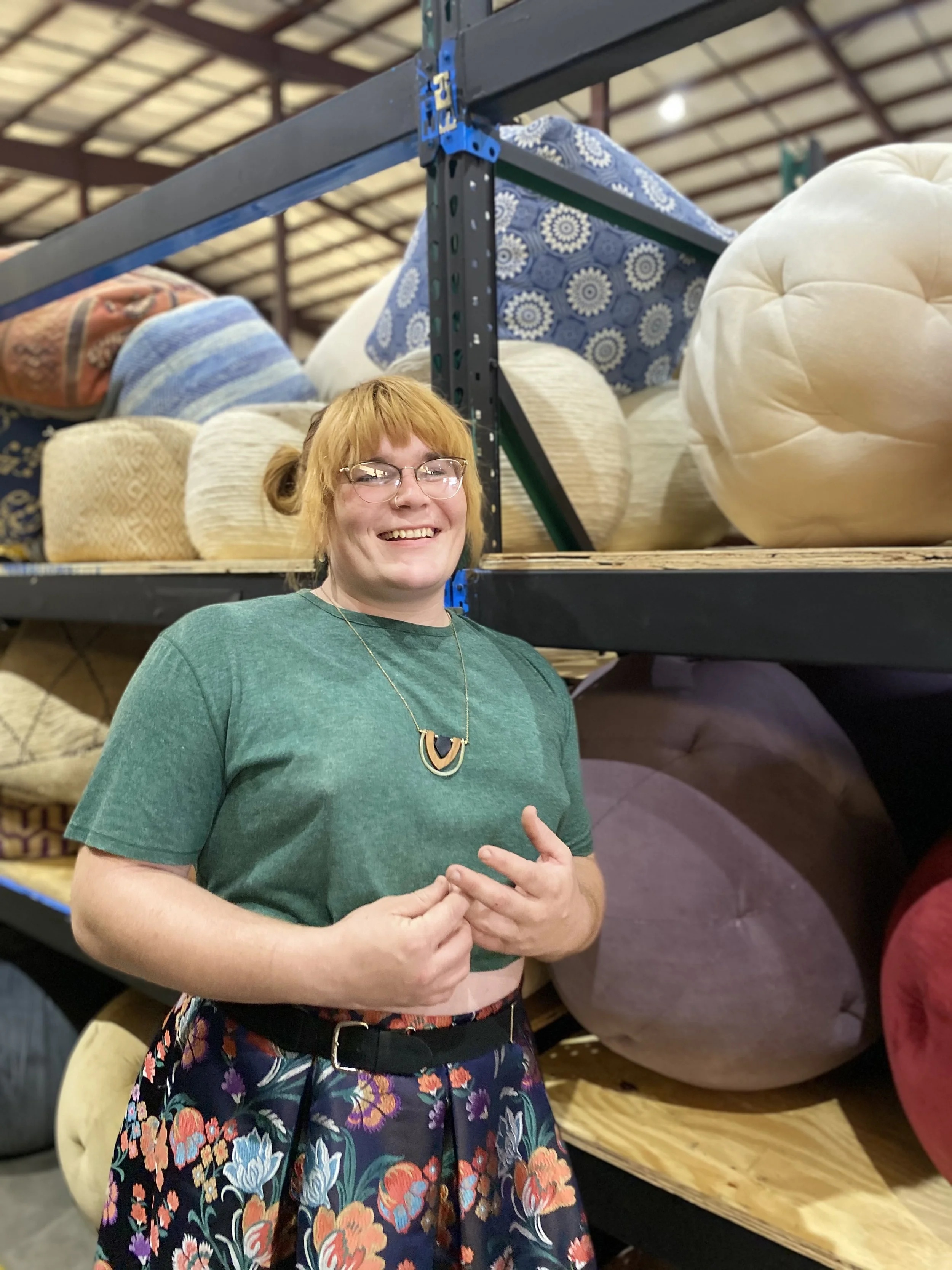 A woman with short red hair and glasses smiling at the camera, standing in front of a display of assorted mattresses in a store.