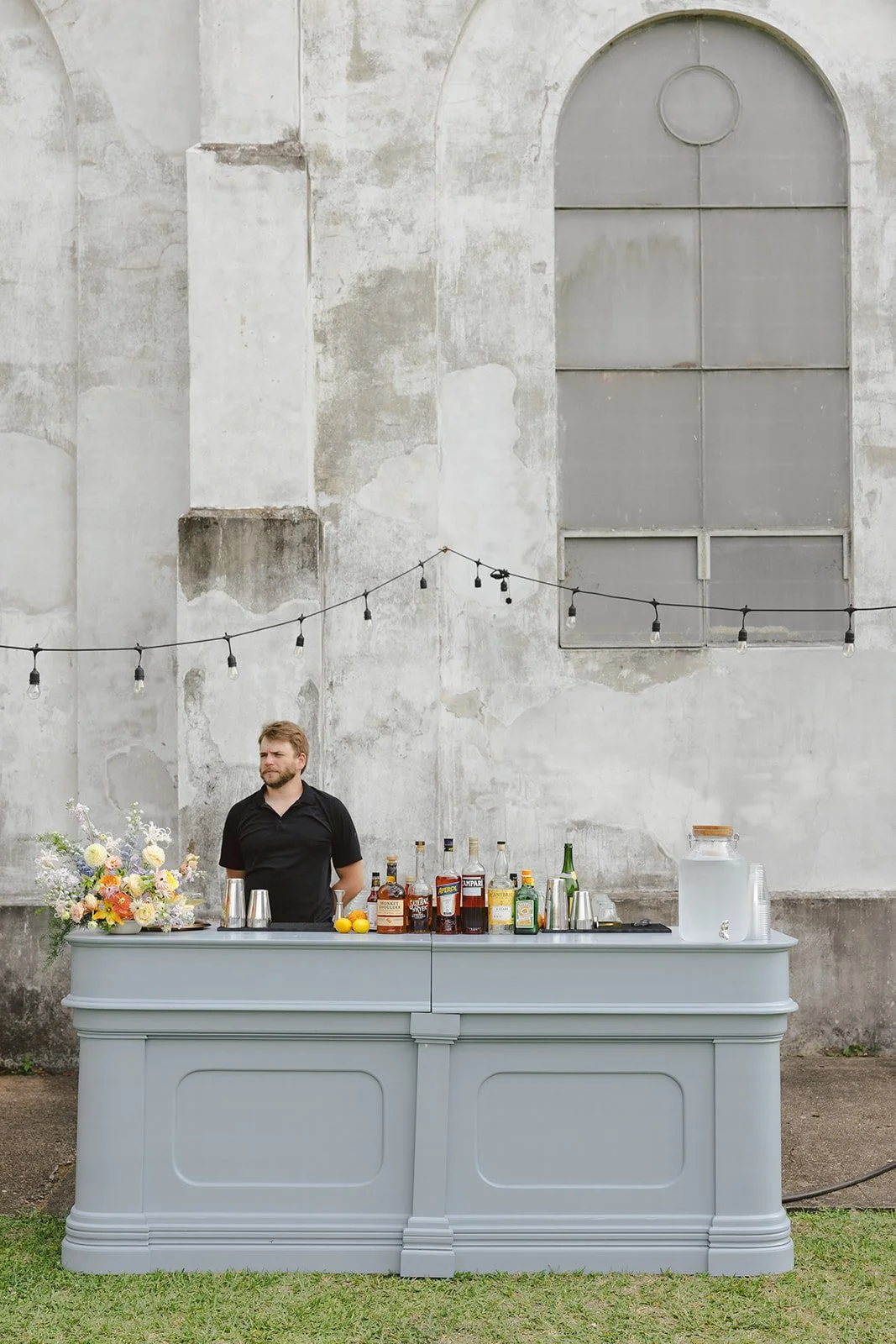 A bartender standing behind a white outdoor bar with bottles of alcohol, lemons, and floral arrangements, in front of a weathered concrete wall with a large arched window and string lights overhead.