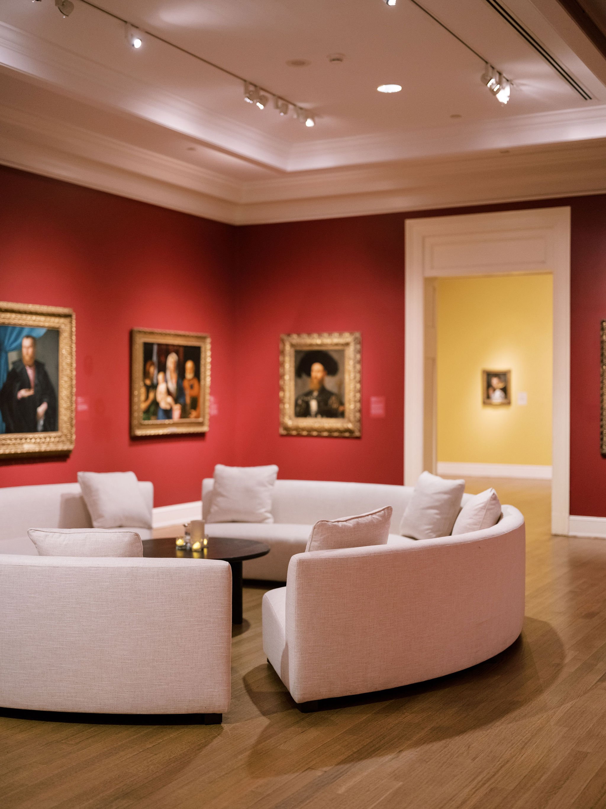 A cozy seating area in an art gallery or museum with a white curved sofa with white pillows surrounding a round black coffee table with candles. The wall behind features framed classical portraits and a doorway leading to another room painted yellow.