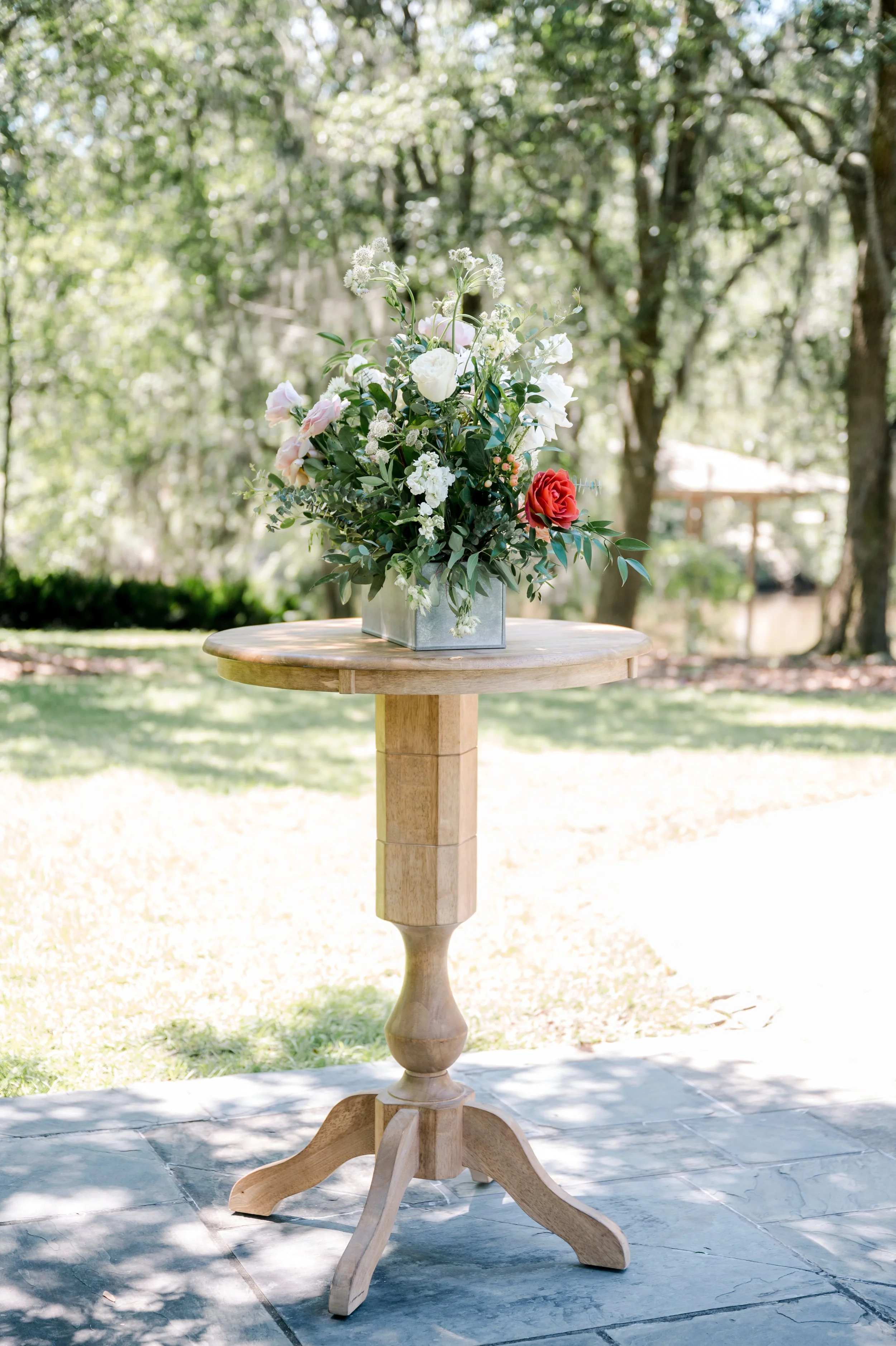 A wooden round table with a flower arrangement in a metal vase, set outdoors with trees and sunlight in the background.