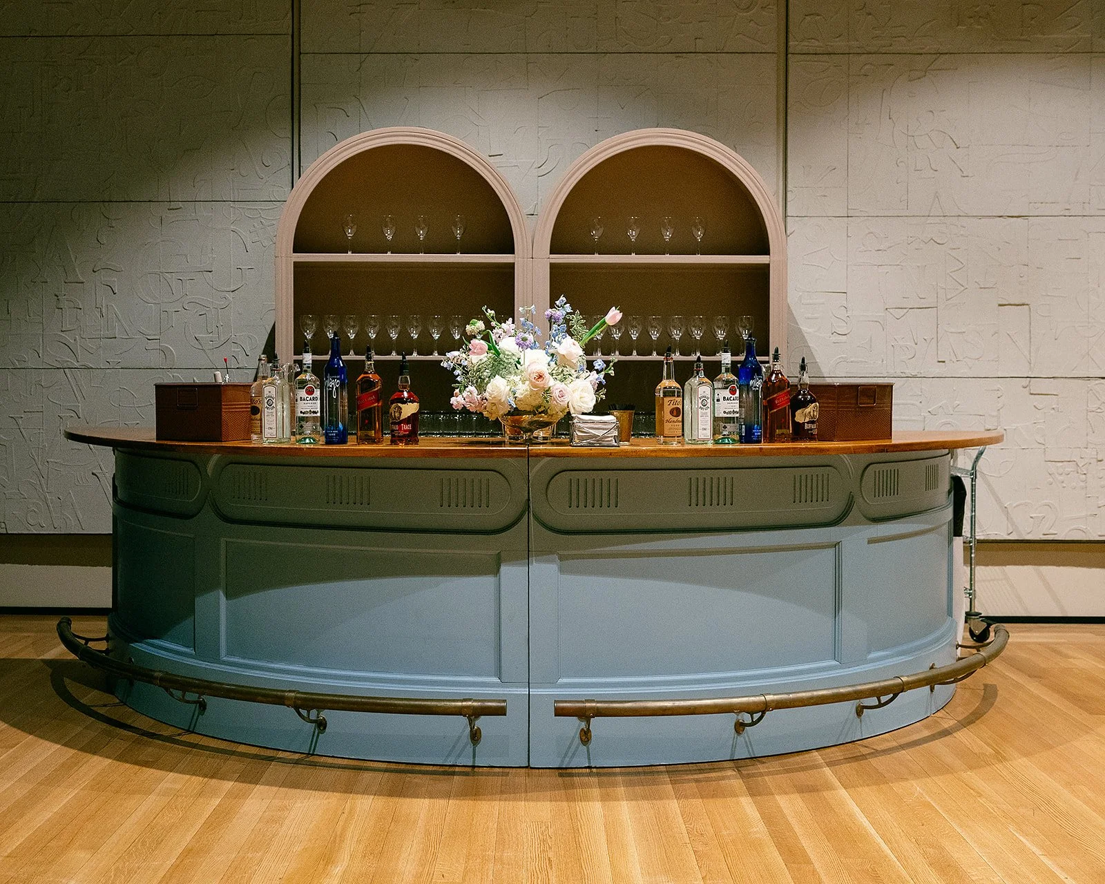 Bar counter with a light blue base, topped with various bottles of alcohol, a box, and a floral arrangement in the center. There are glasses on a shelf behind the counter and a white textured wall in the background.