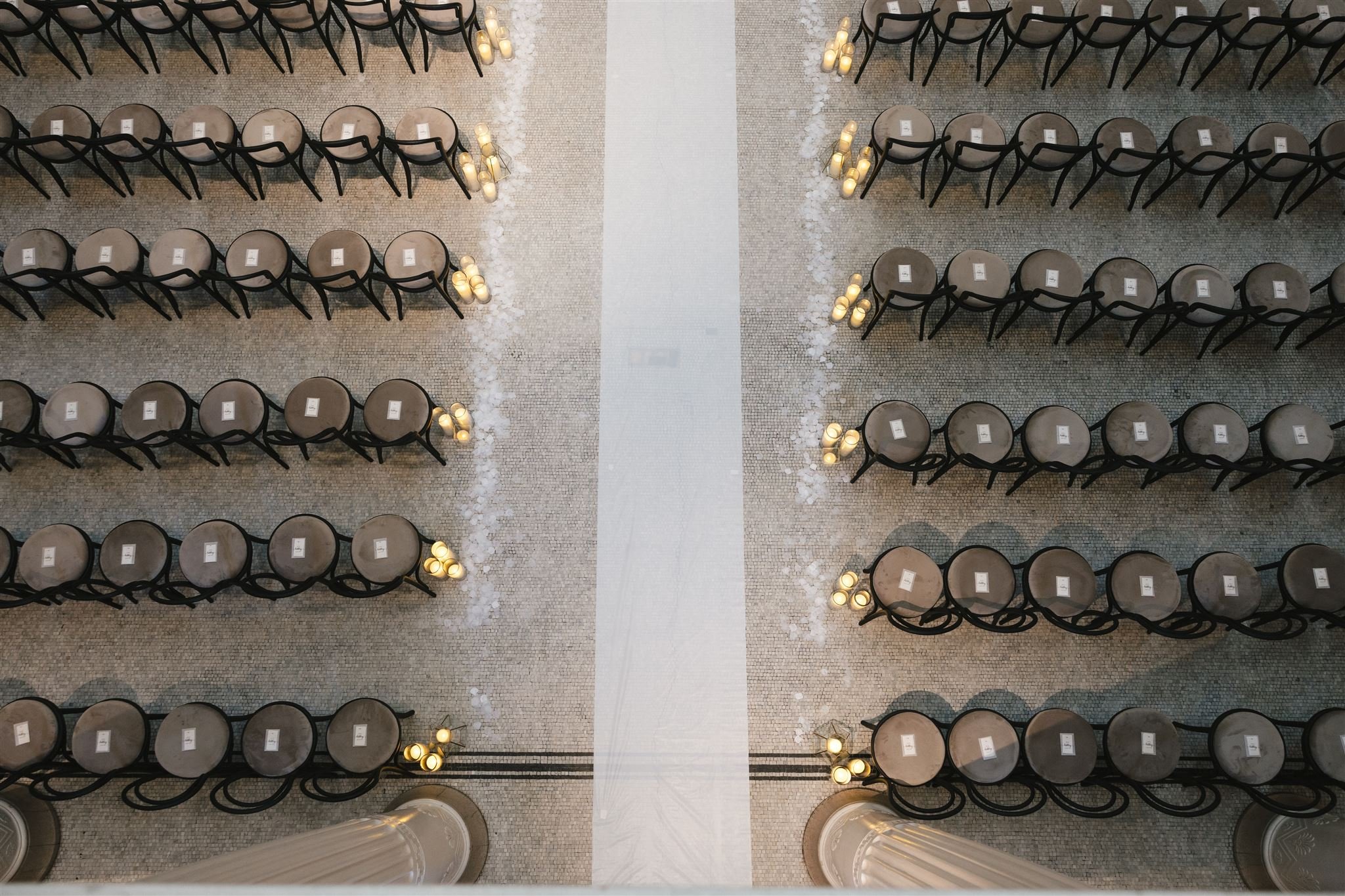 Overhead view of rows of beige chairs with black frames arranged in a large hall, with candles on the floor and a white aisle in the middle.
