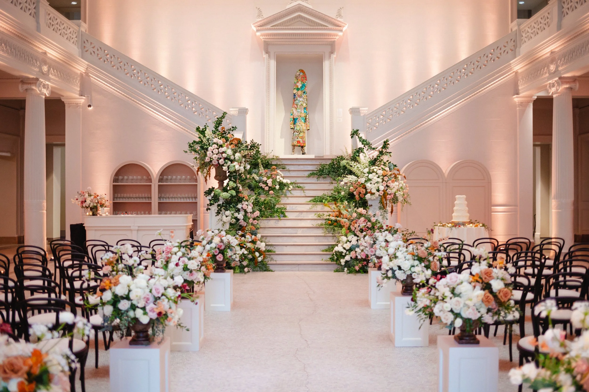 Elegant wedding ceremony setup in a grand indoor hall with floral arrangements along the aisle and staircase, leading to a decorative backdrop with a dress on display.