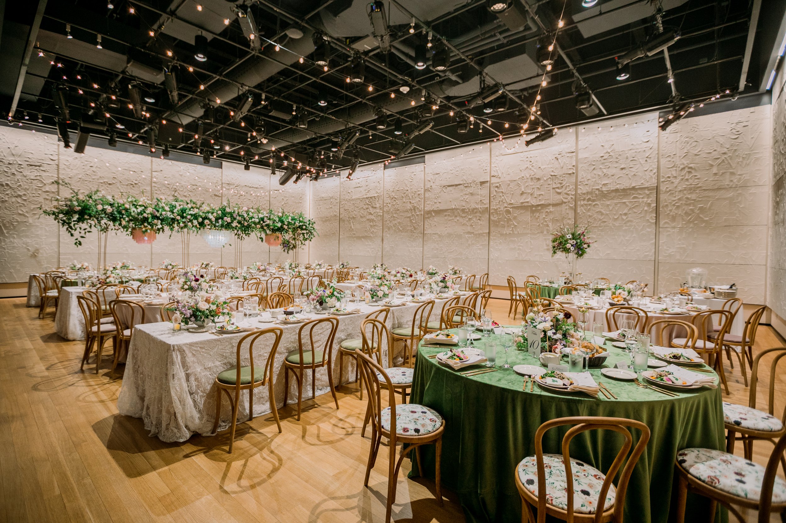 An indoor event space decorated for a celebration with round tables covered in white and green tablecloths, floral centerpieces, and elegant chairs. String lights are hung from the black ceiling with extensive lighting and sound equipment. The wall h