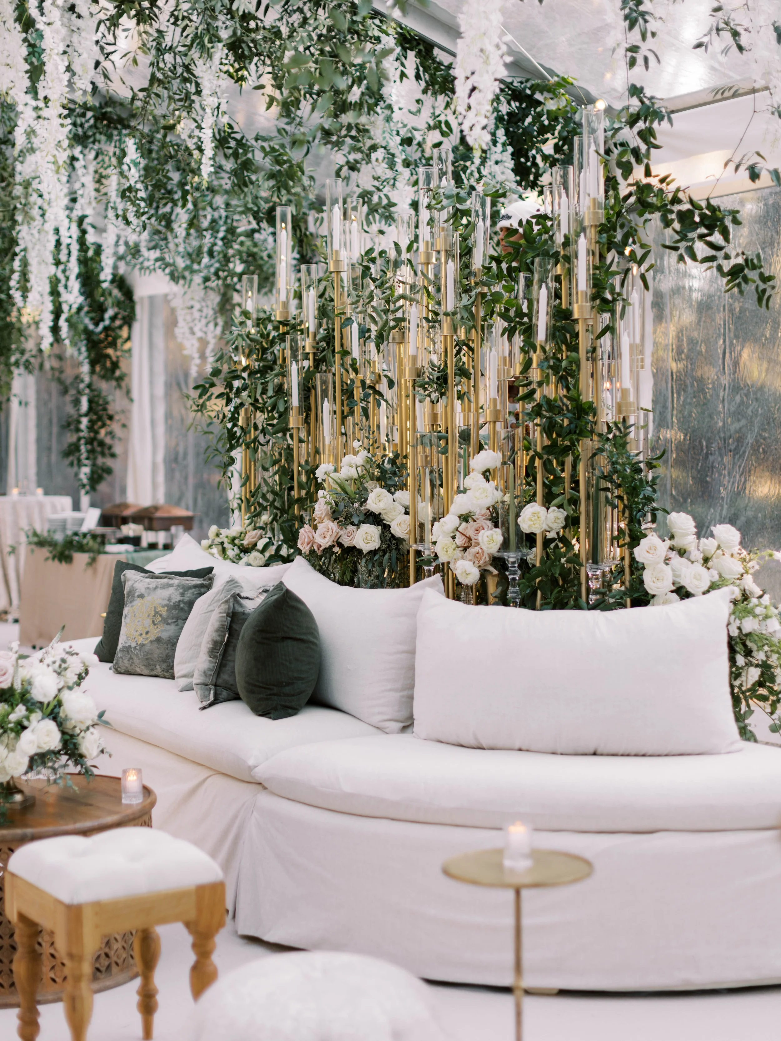Luxurious indoor event setup with a white sofa adorned with dark and light-colored pillows, surrounded by elegant floral arrangements, greenery, and tall glass candle holders.