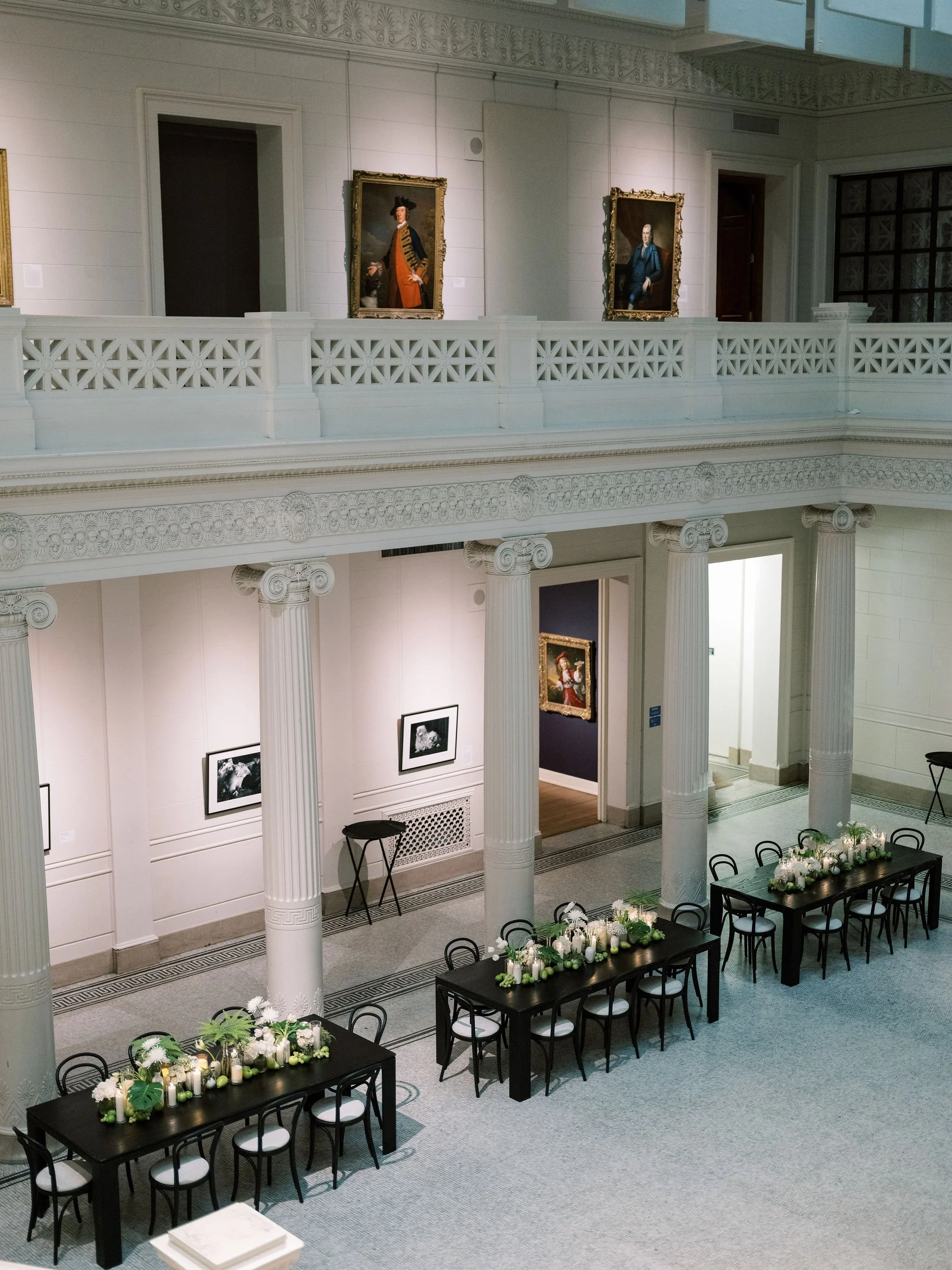 Elegant room with white walls and columns, decorated for an event, featuring tables with floral arrangements and candles.