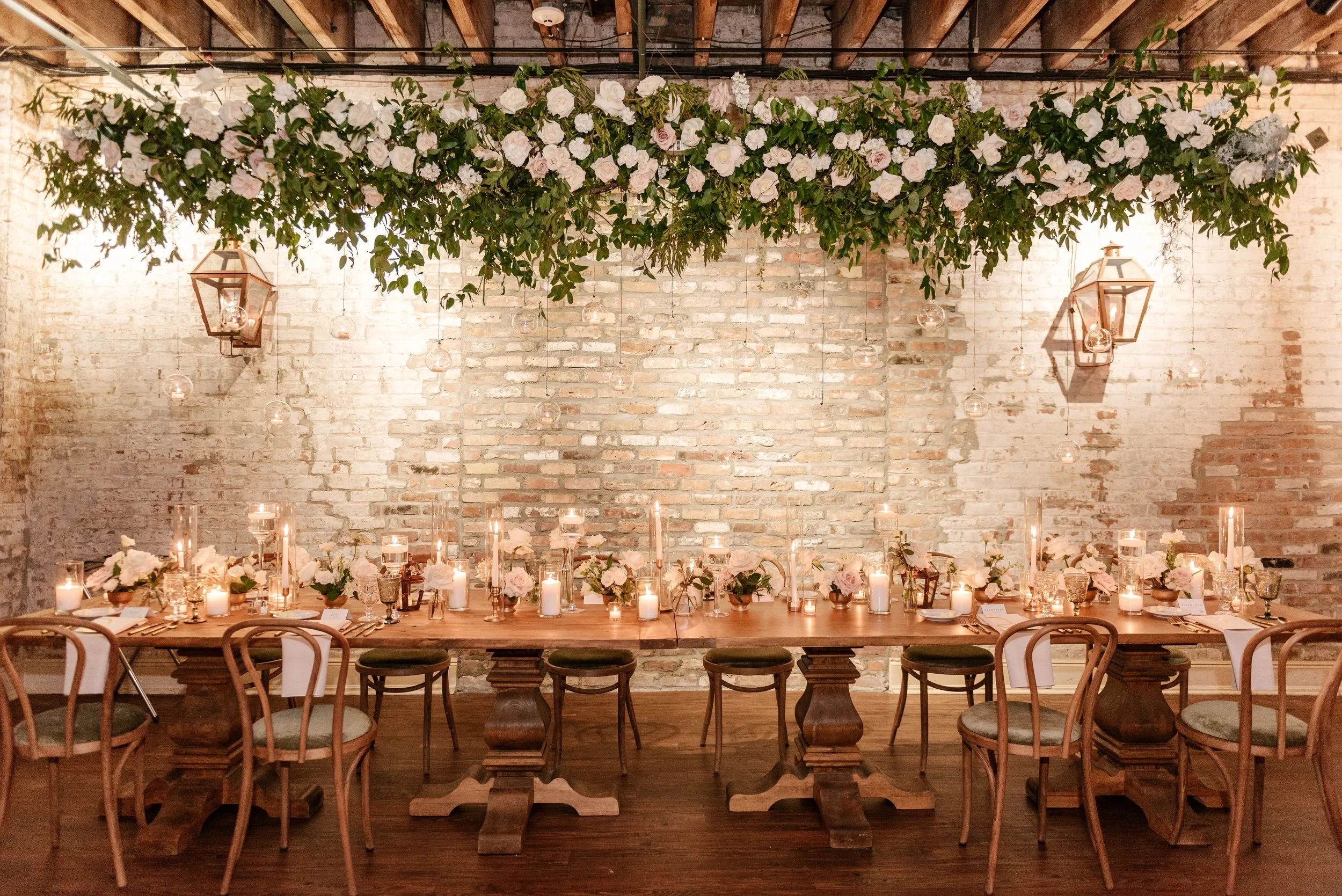 A large wooden table set for a gathering or wedding with white flowers and candles, against an exposed brick wall. Hanging above the table is a decorative greenery installation with white roses and lantern-style lights.