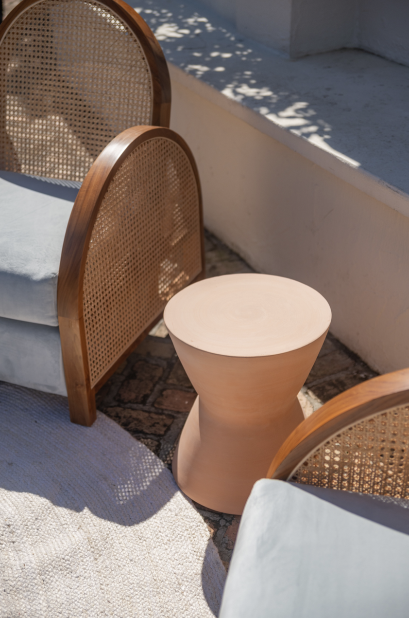 Two wooden chairs with woven cane backs and a light-colored wooden side table, set on a light rug in an outdoor area with white walls and sunlight casting shadows.