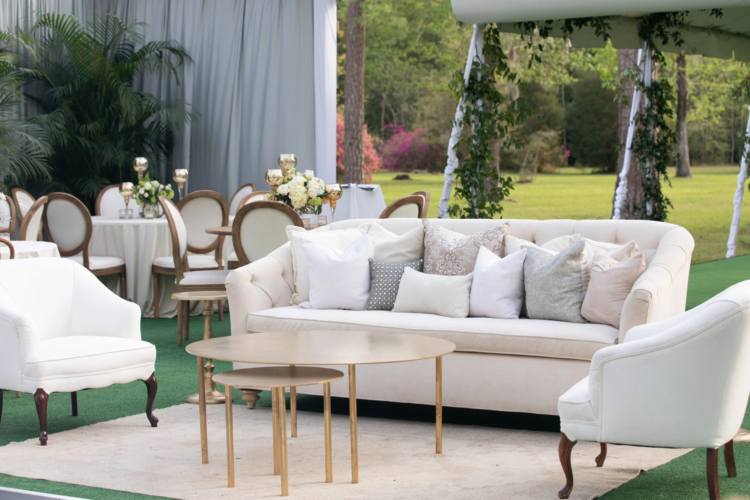 An outdoor wedding or event setup with a white sofa, cream and patterned pillows, wooden tables, a round dining table with floral arrangements, and a decorated pavilion with greenery, set in a grassy area with trees in the background.