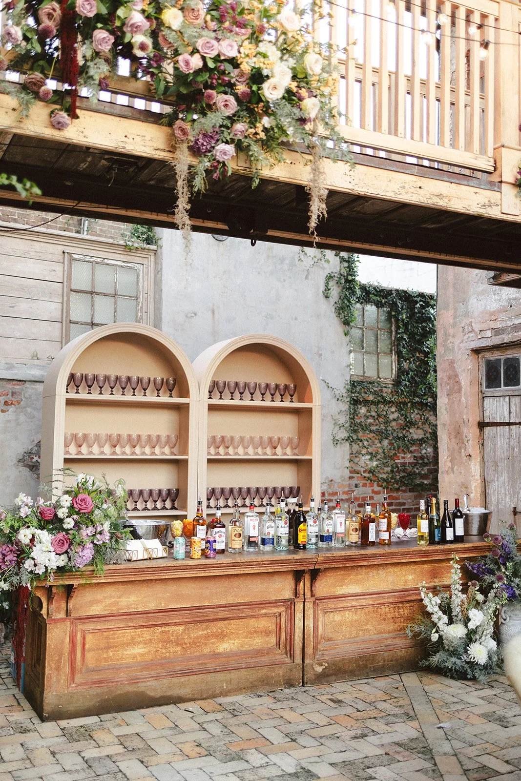 A rustic outdoor bar setup with bottles of alcohol on a wooden counter, surrounded by floral arrangements, with a decorative wooden cabinet and brick wall in the background.