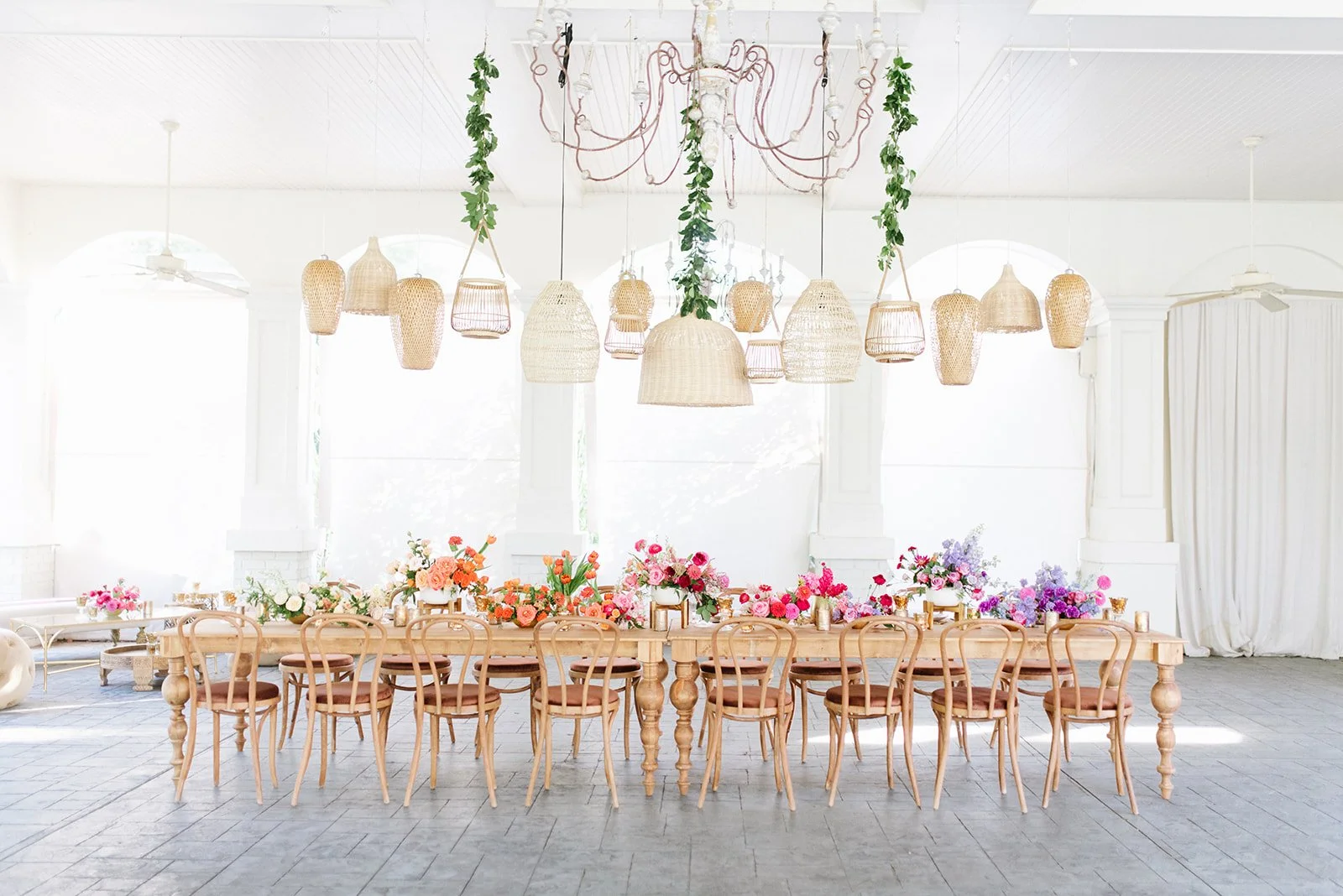 A long wooden table decorated with colorful flower bouquets, surrounded by wooden chairs in a bright, white room with large windows and hanging rattan and wicker pendant lights, creating a soft, elegant atmosphere.