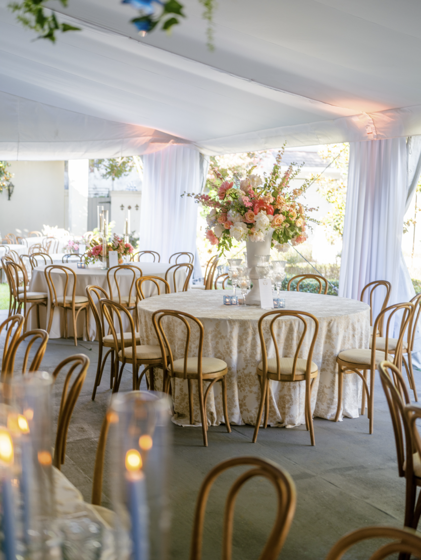 A decorated event tent with round tables covered in cream tablecloths, set with floral centerpieces and glassware, surrounded by wooden chairs with cream cushions, and soft lighting creating a warm atmosphere.